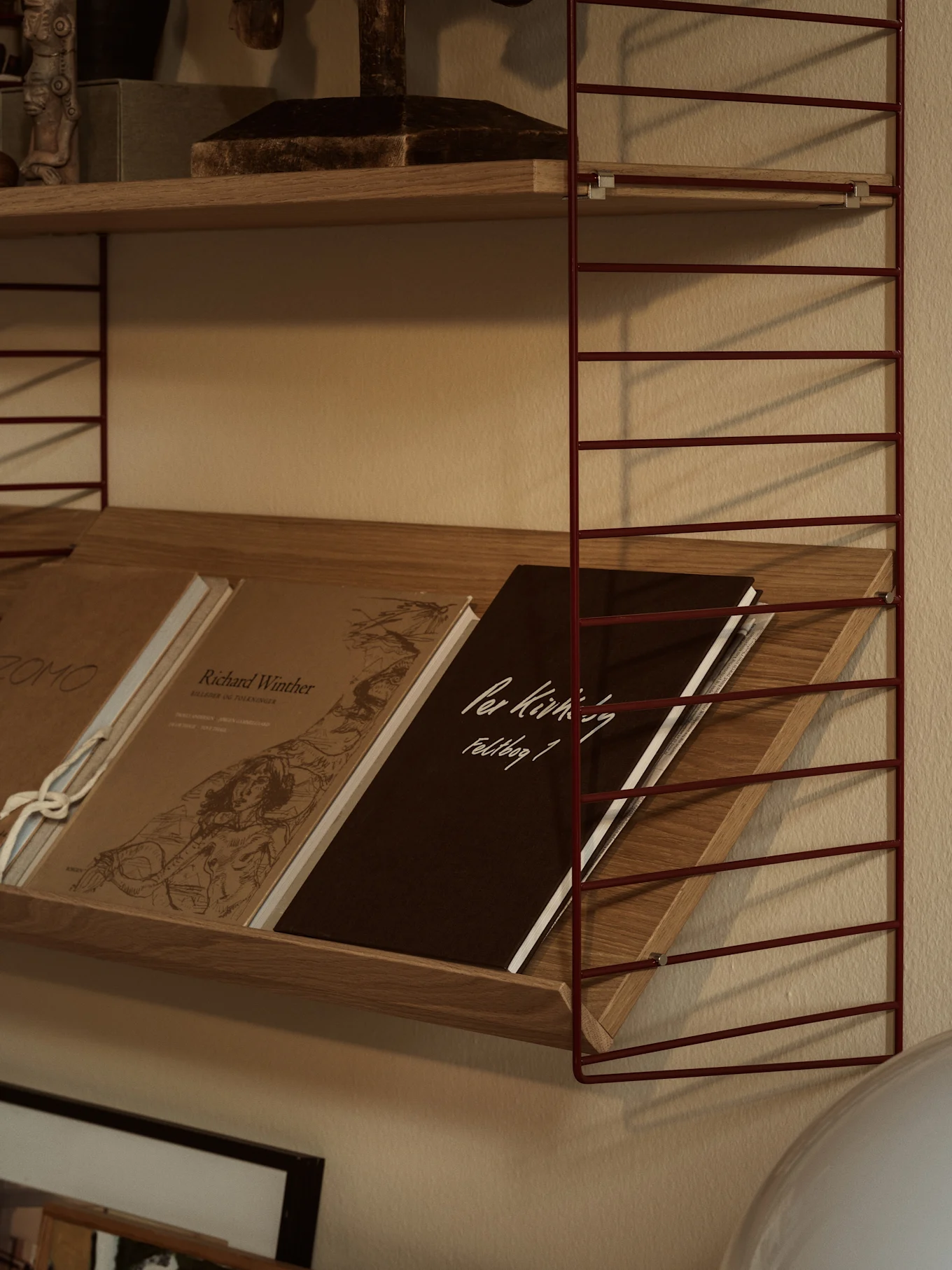 A red metal wire shelving unit with wooden shelves. The lower shelf holds angled books, including a dark brown one with 'Per Kirkeby Feldbog 1'.