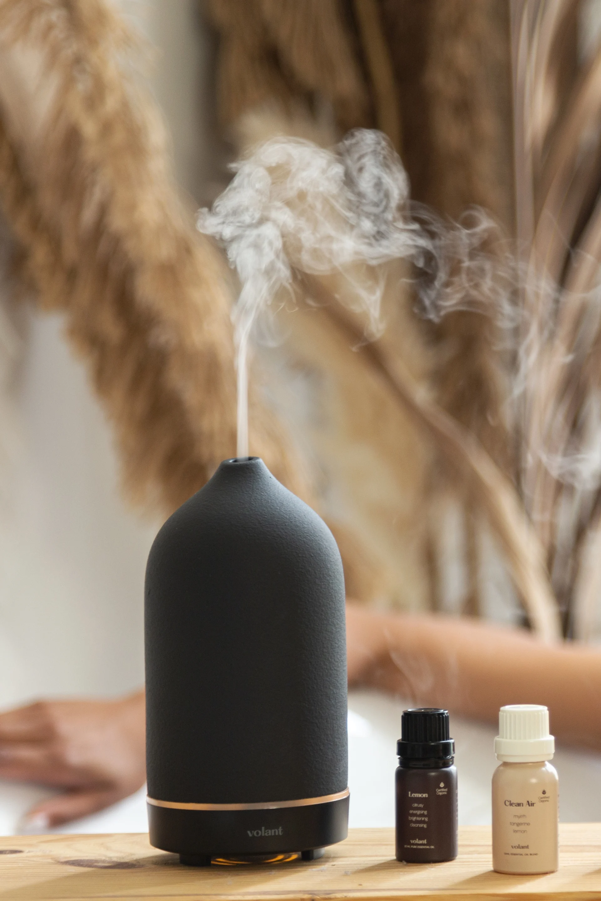 A black essential oil diffuser emits mist, flanked by 'Lemon' and 'Clean Air' essential oil bottles on a wooden surface.