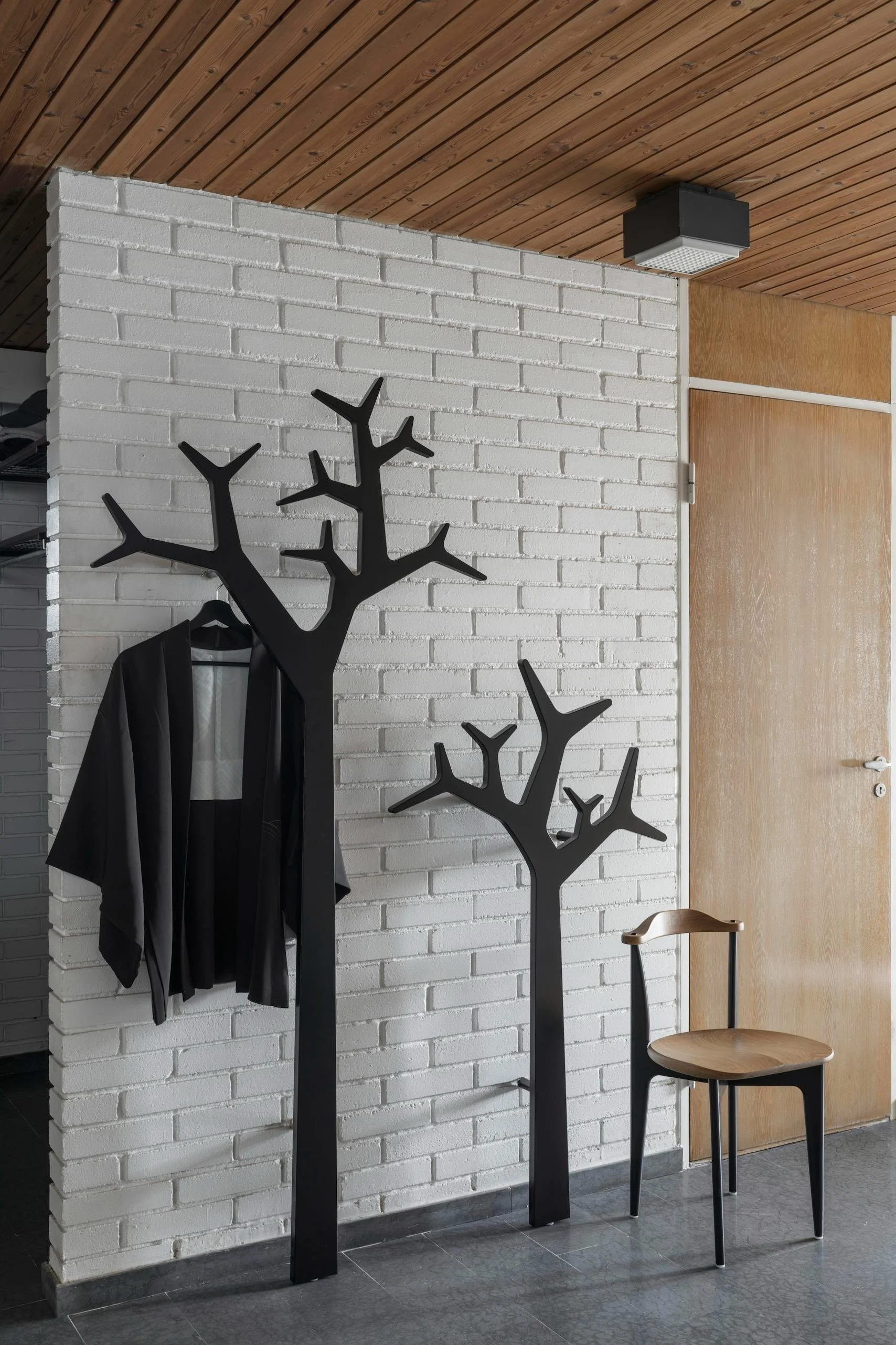 Two black tree-shaped coat stands, one with a black jacket, against a white brick wall in a room with a wood ceiling.