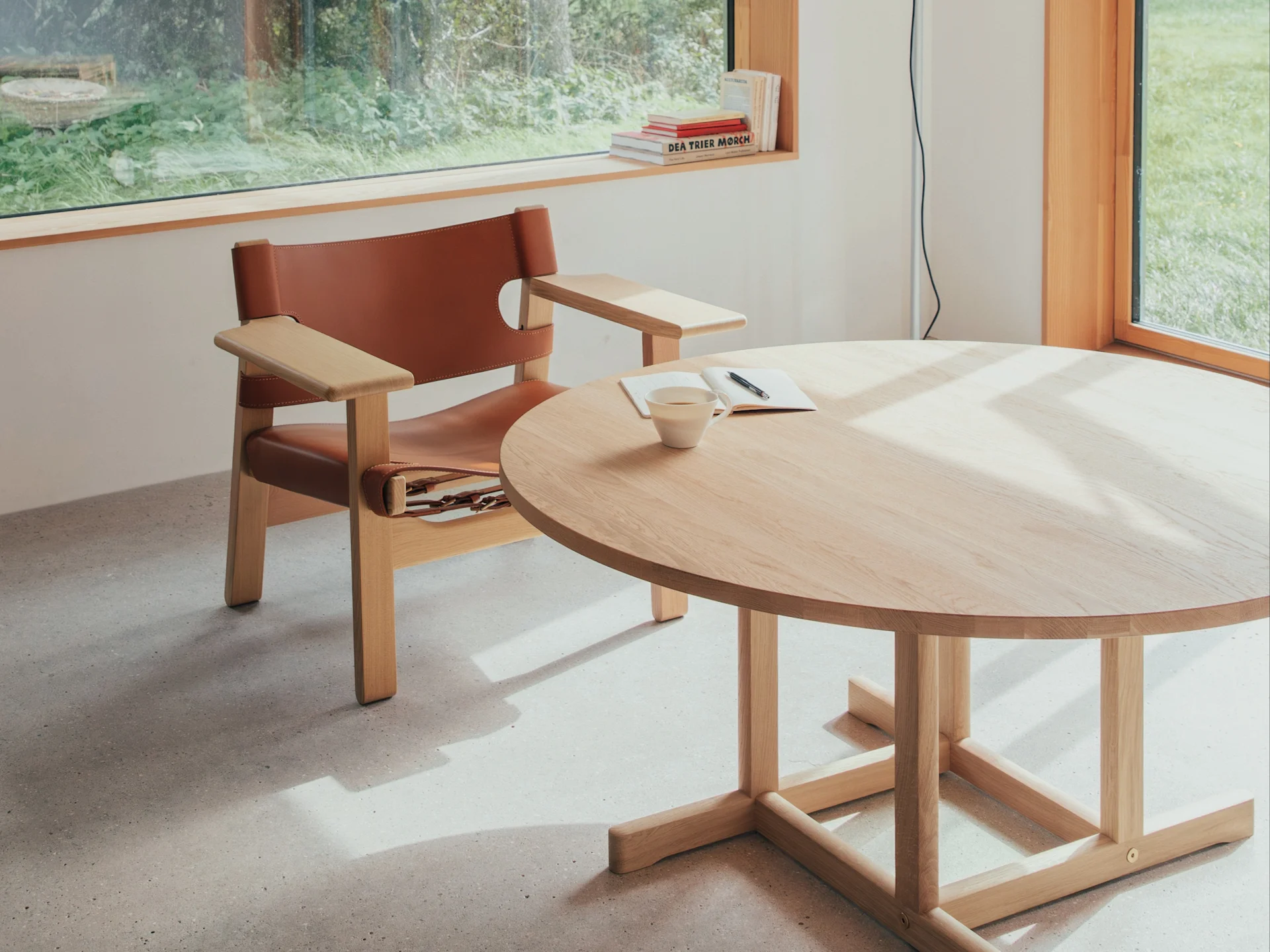 Bright room with a brown leather armchair, a round wood table with a cup and notebook, sunlit by a window overlooking greenery.