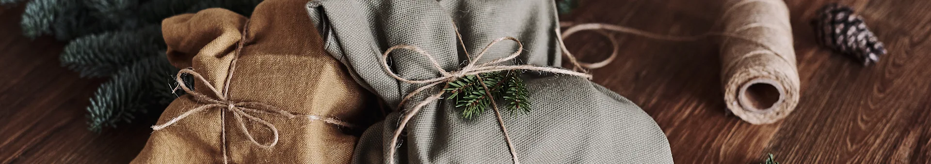 Two fabric-wrapped gifts, one brown and one green, tied with twine and decorated with a pine branch on a wooden surface.