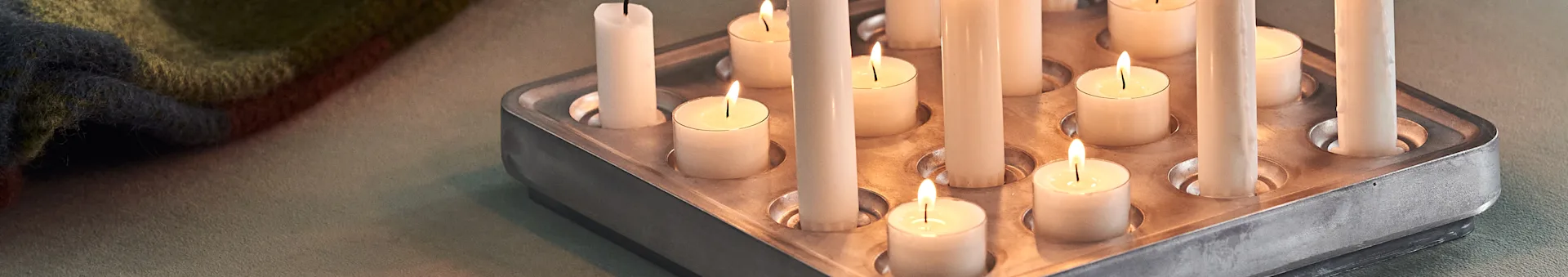Close-up of a metal tray with several lit and unlit white candles, a patterned blanket in the background.