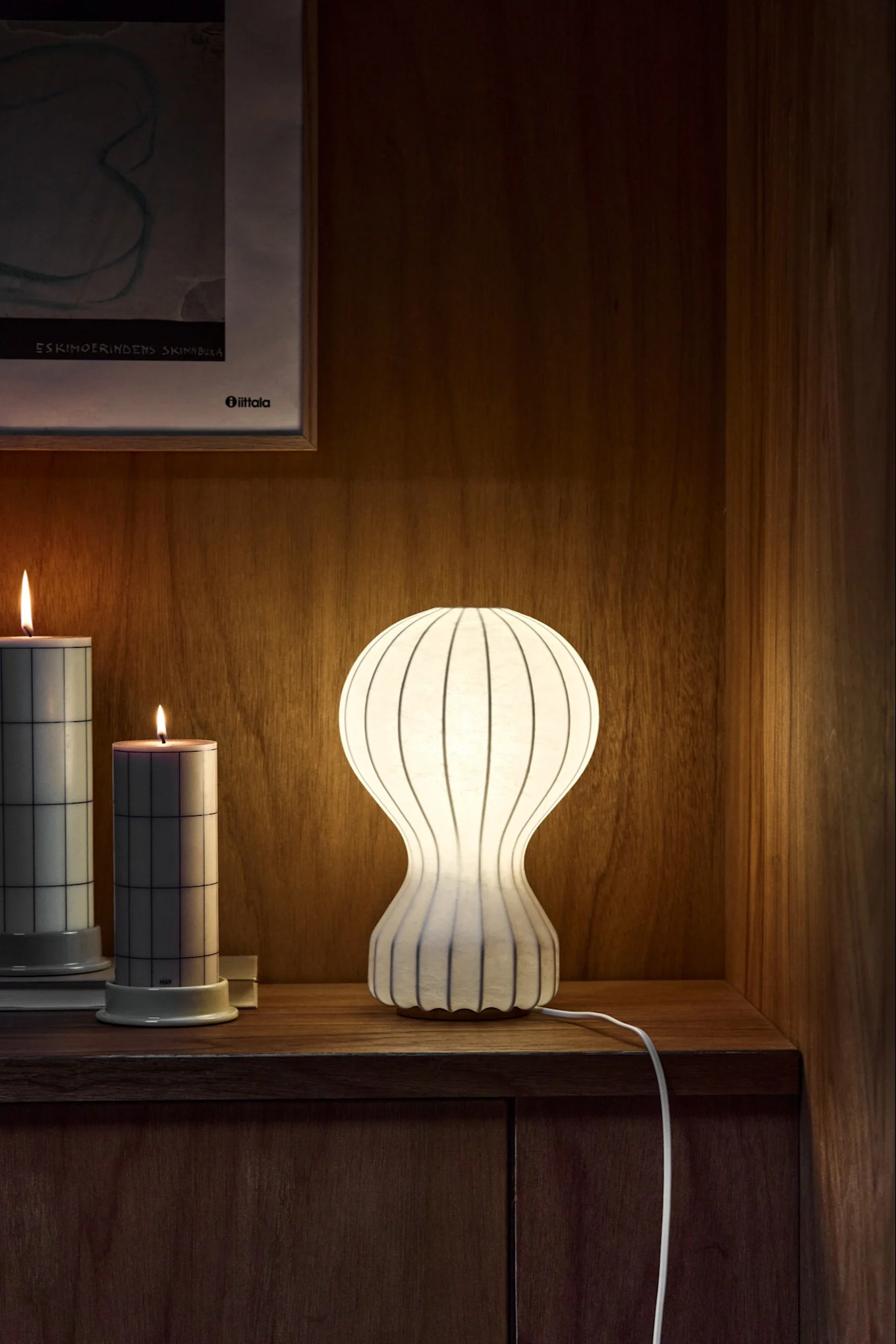An hourglass-shaped ribbed white table lamp and two grid-patterned lit candles on a wooden surface.
