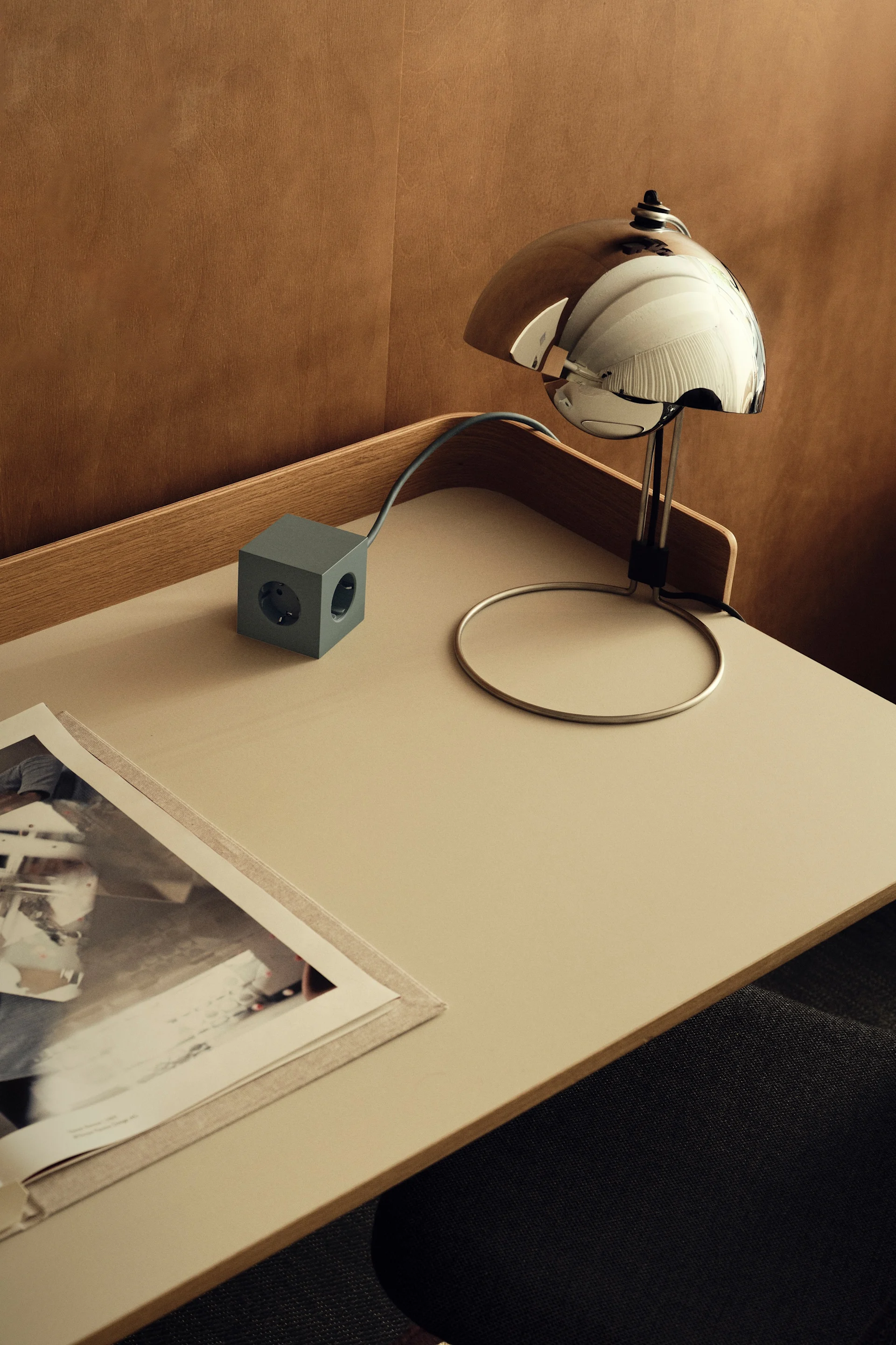 A minimalist desk setup with a chrome dome lamp, a grey cube power strip, and an open book on a light-colored surface.