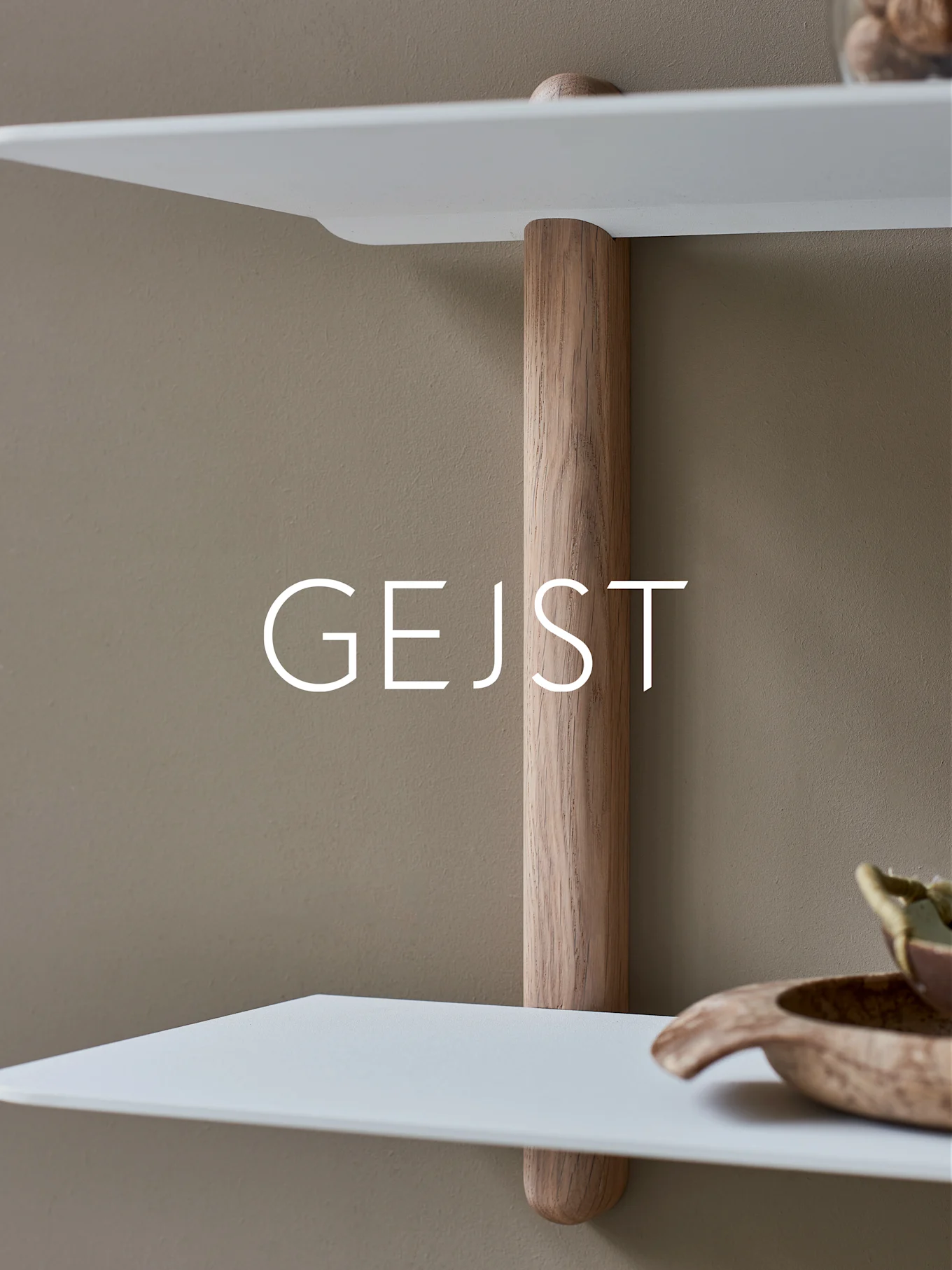 Two white, rounded shelves connected by a vertical light wood support pole against a beige wall. A wooden bowl is on the lower shelf.