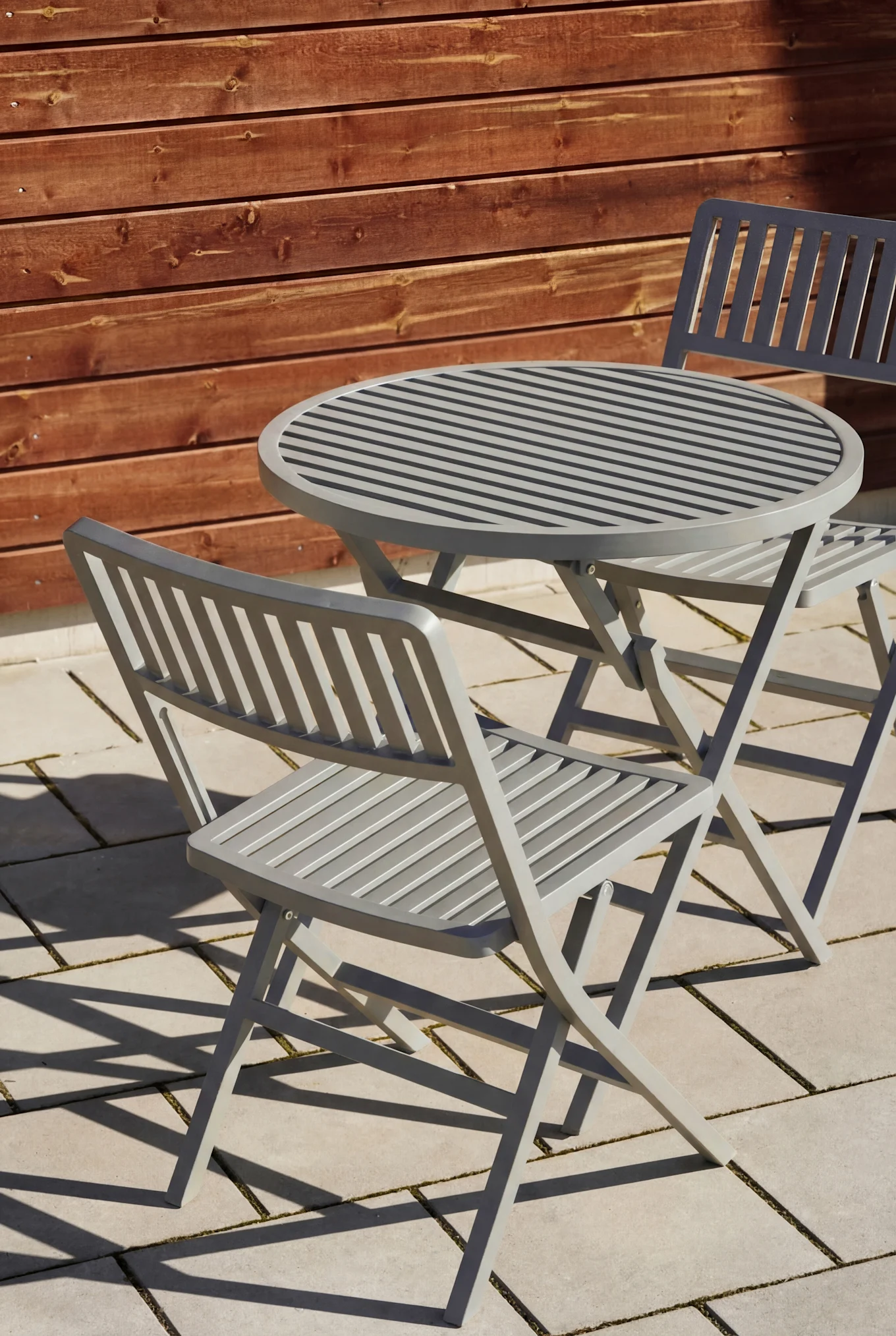 Light gray slatted outdoor table and two chairs on a patio with a wooden fence in the background.