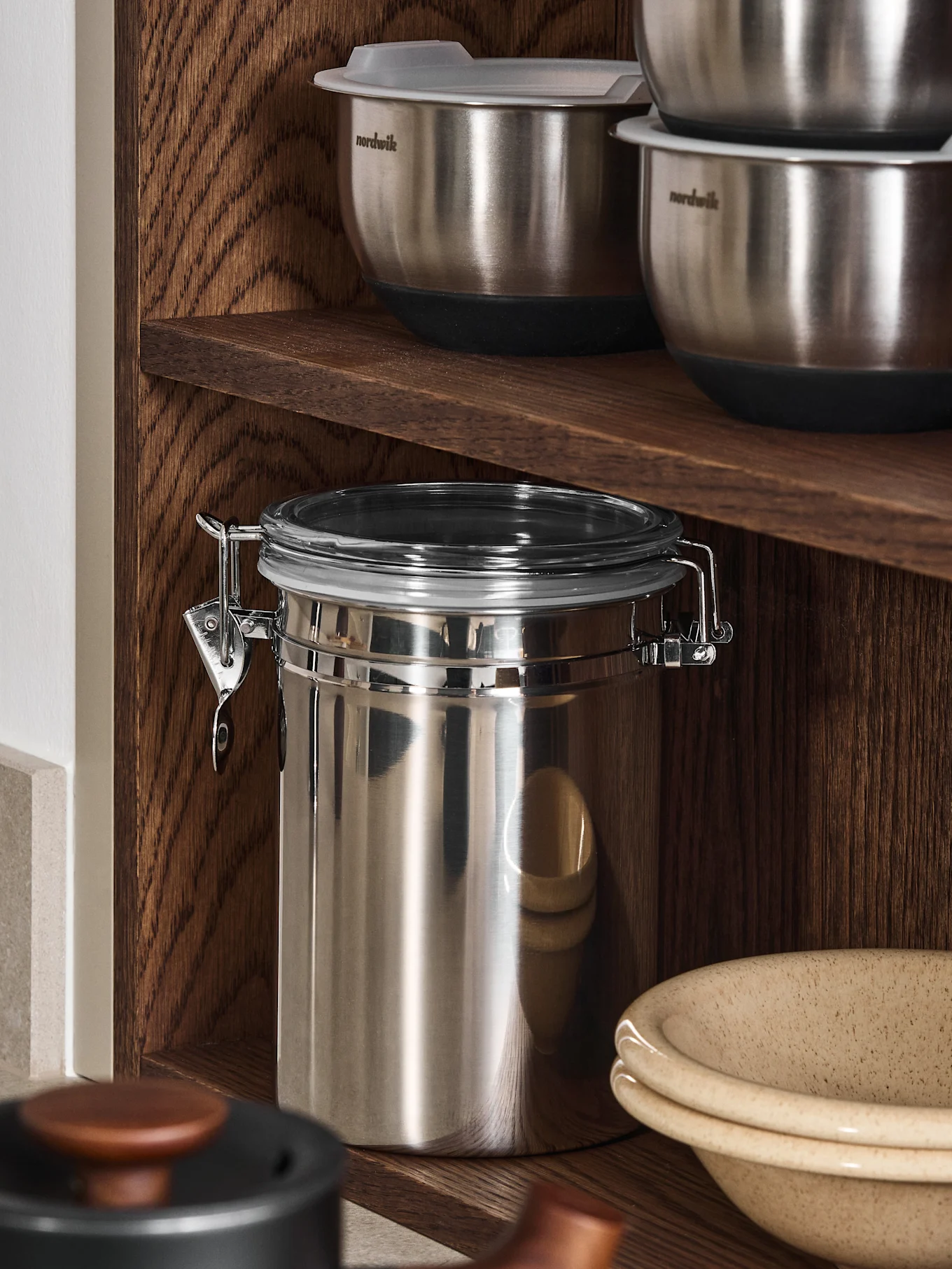 Stainless steel mixing bowls and a large storage jar on dark wooden shelves, with a ceramic bowl below.
