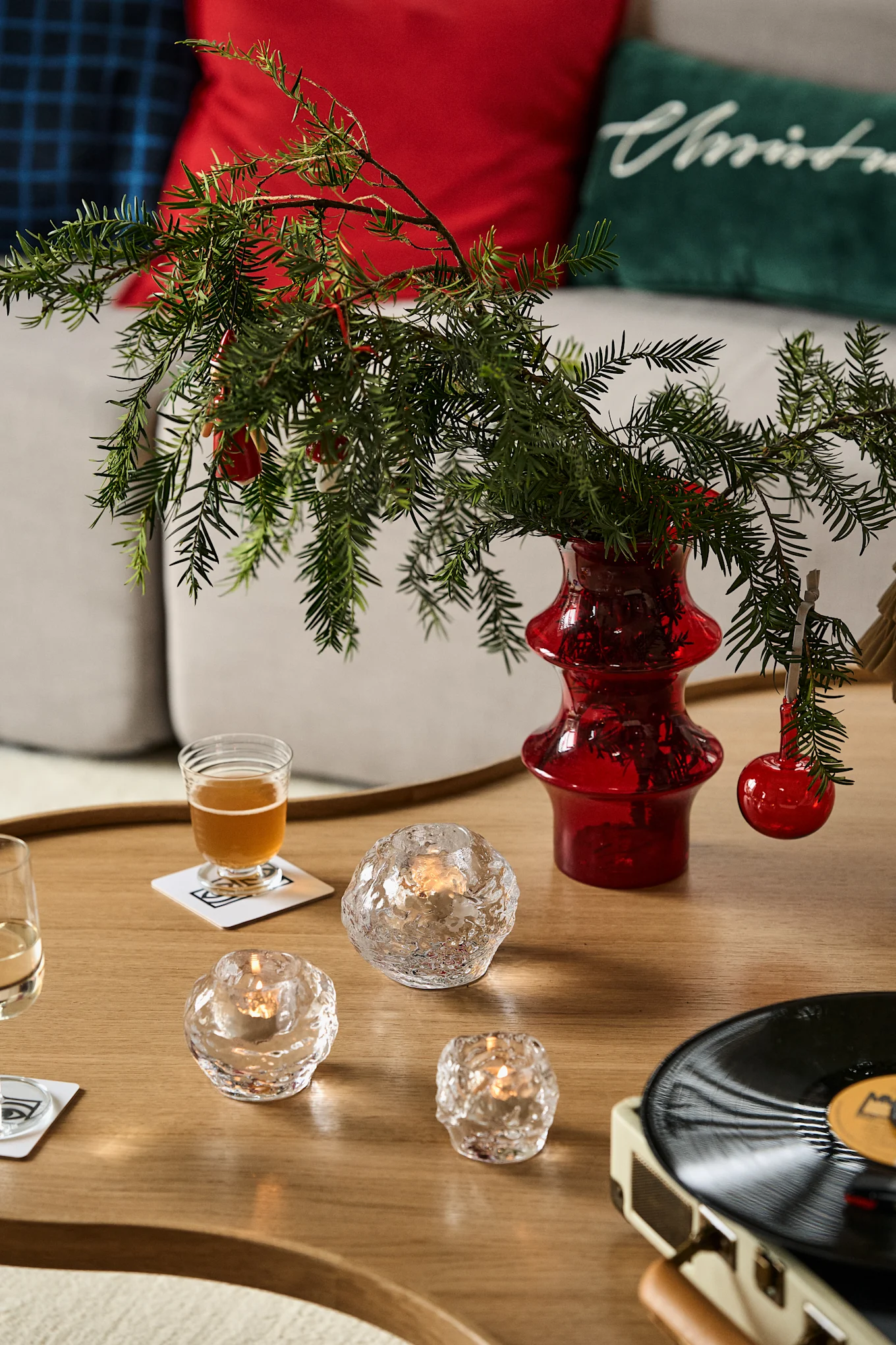A coffee table with holiday decor: red vase with evergreen branches, candles, drinks, and a record player.