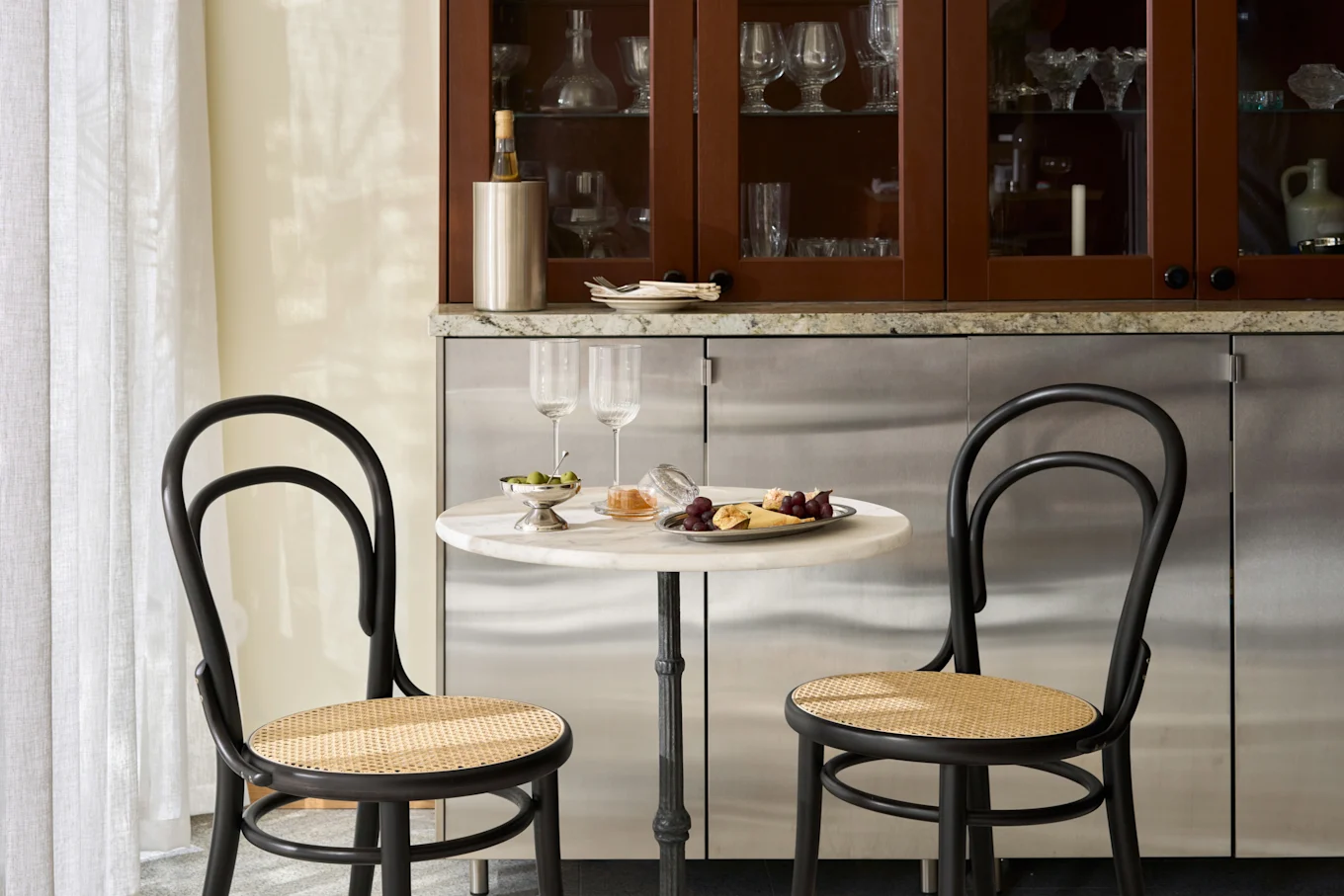 Cozy dining nook with marble table, two black cane chairs, a platter of cheese and fruit, olives, and wine glasses.