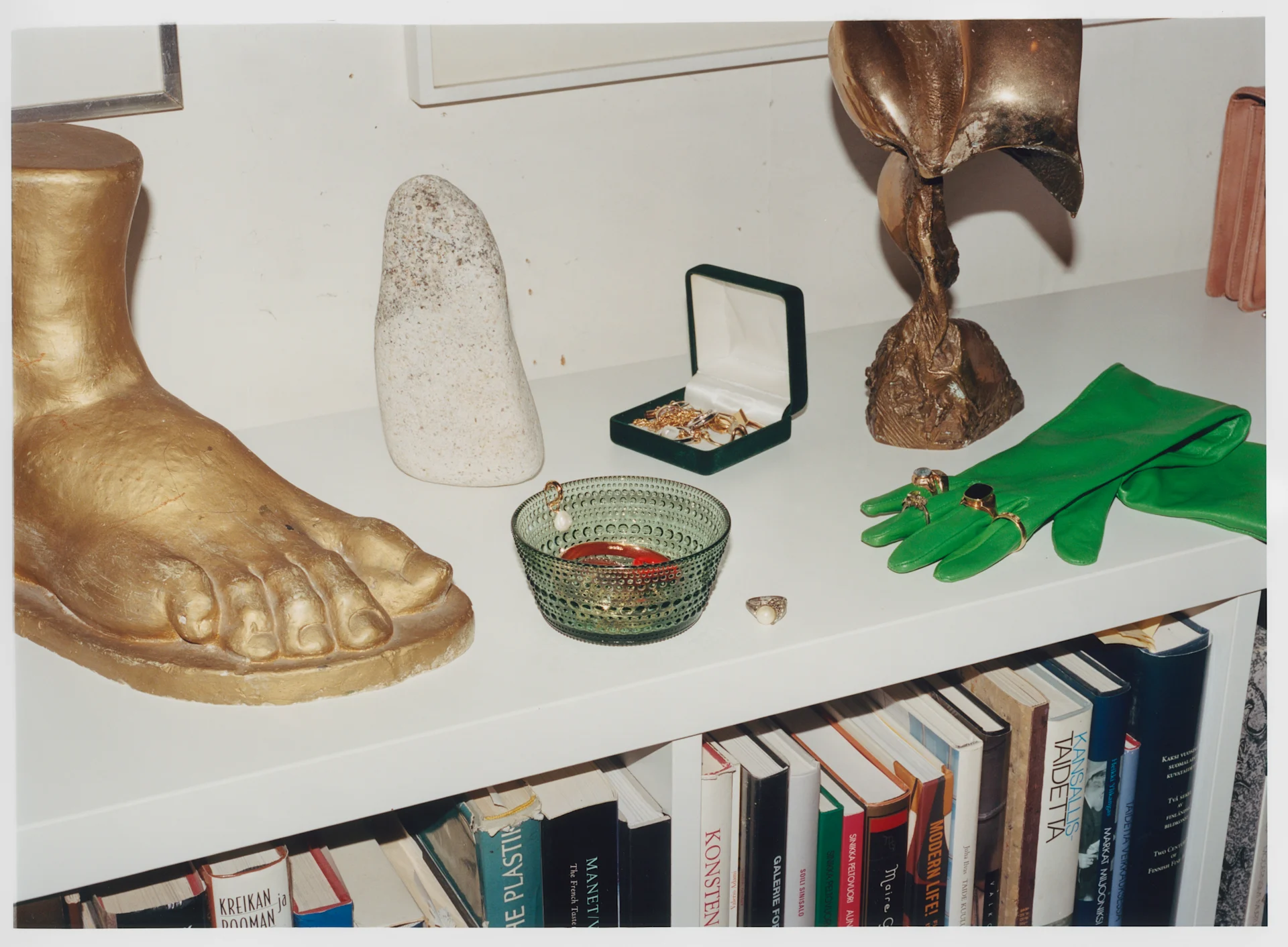 A white shelf displays a golden foot sculpture, a stone, an open green jewelry box, a green glass bowl, and green gloves with rings. Books are below.