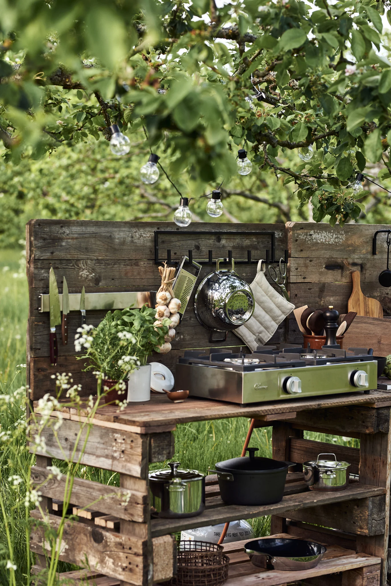 An outdoor kitchen made from wooden pallets with a green two-burner gas stove, hanging utensils, and potted herbs under string lights.