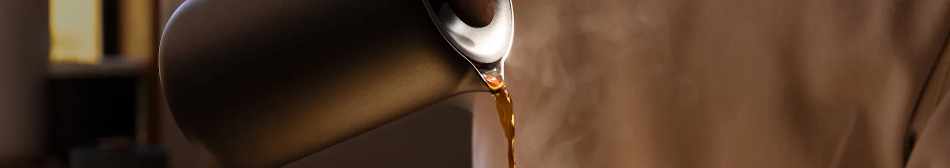 Dark pour-over coffee pot tilted, pouring hot amber liquid with steam rising.