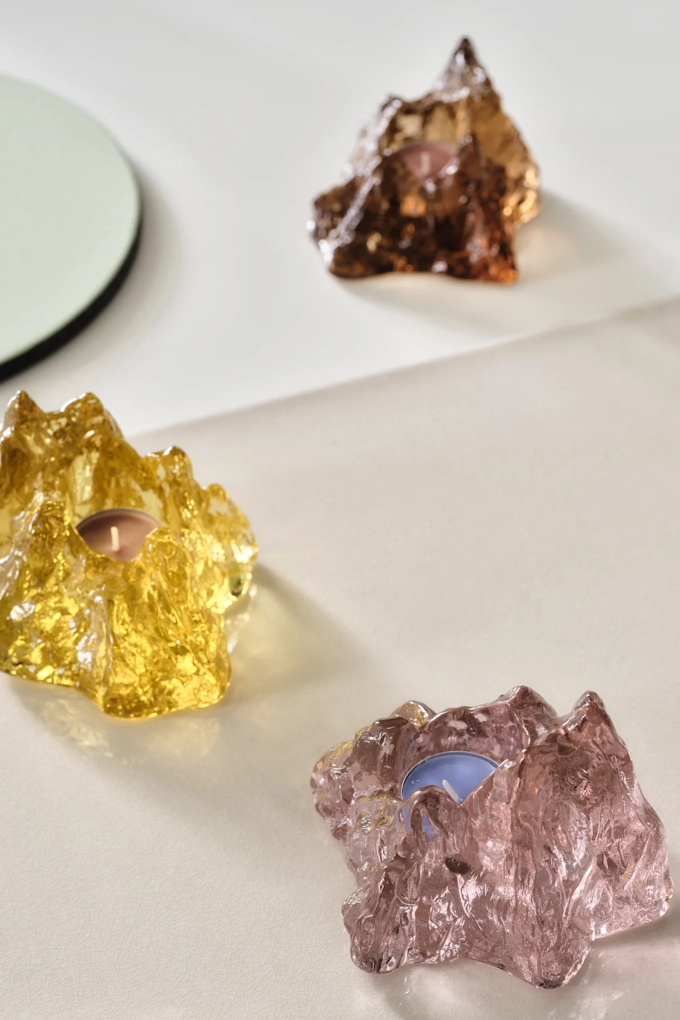 Three textured glass tea light holders in yellow, pink, and amber on a light surface.