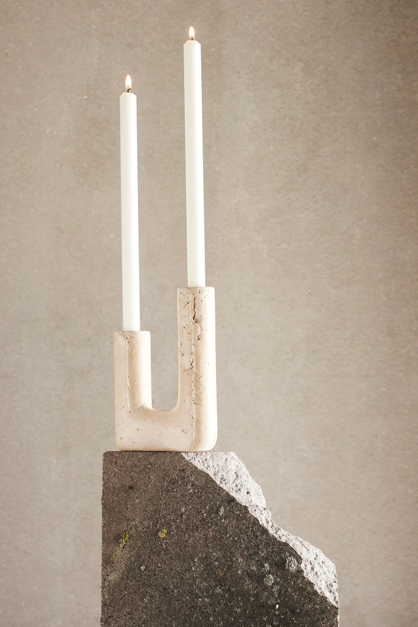 Two lit white candles in a U-shaped light beige stone candle holder, sitting on a dark grey rough stone block against a light beige background.
