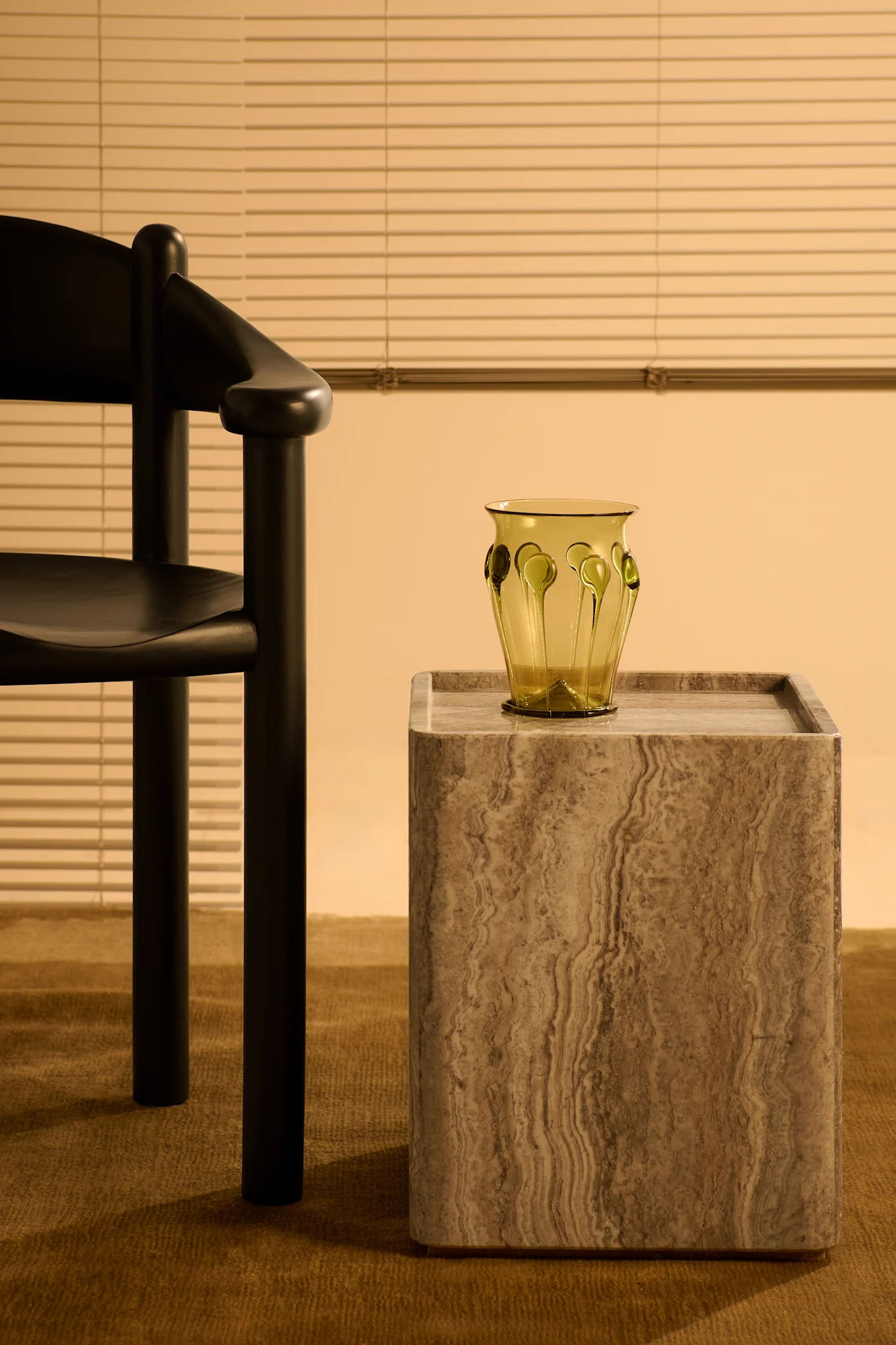 Modern dark wooden chair, stone side table with green glass vase, against a wall with blinds on a brown carpet.