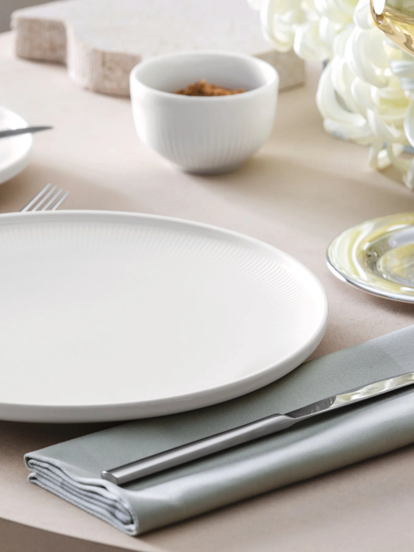 A white ribbed dinner plate, sage green napkin, and silver cutlery set on a table with a small bowl in the background.