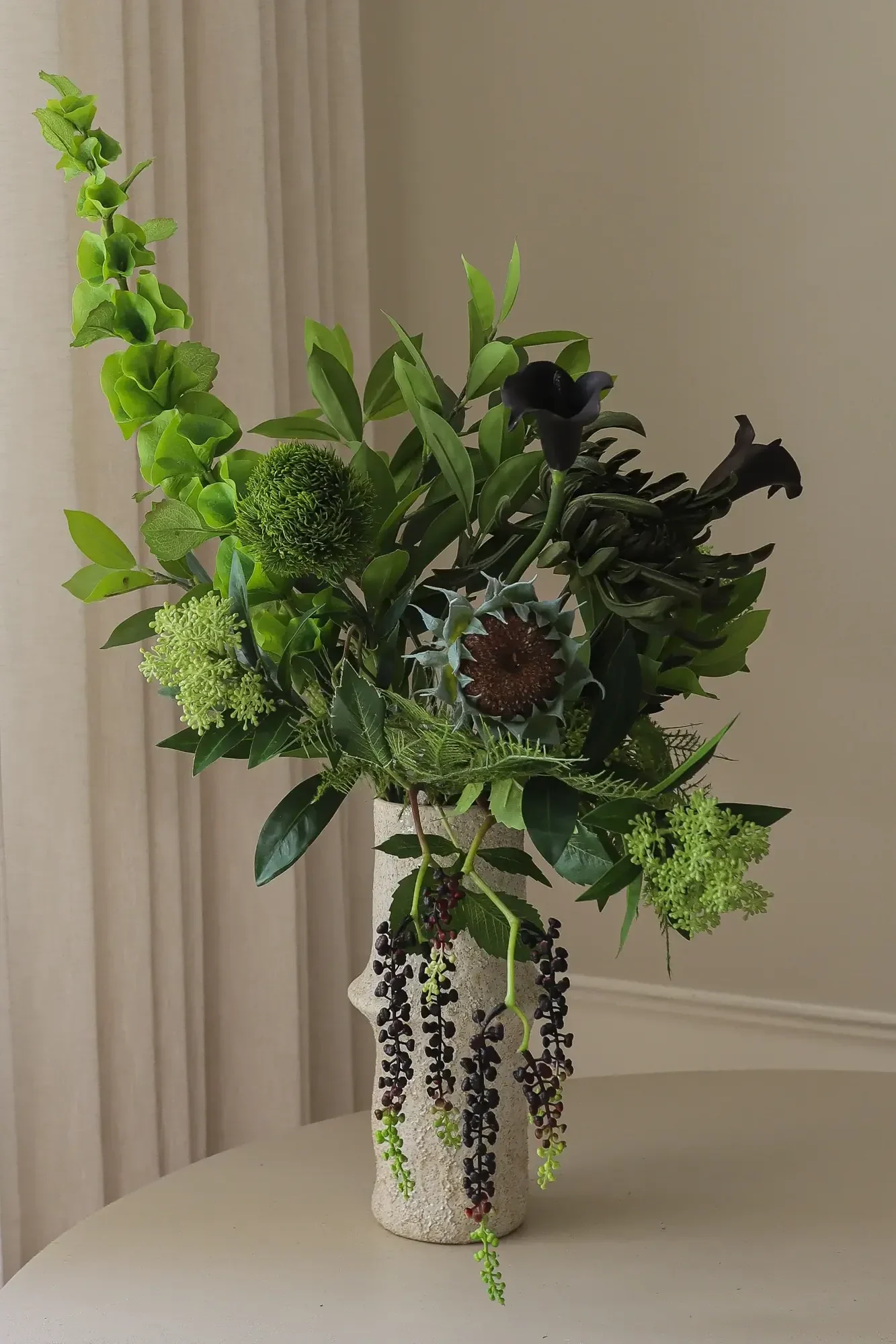 An arrangement of green foliage, dark calla lilies, and hanging berries in a textured vase.