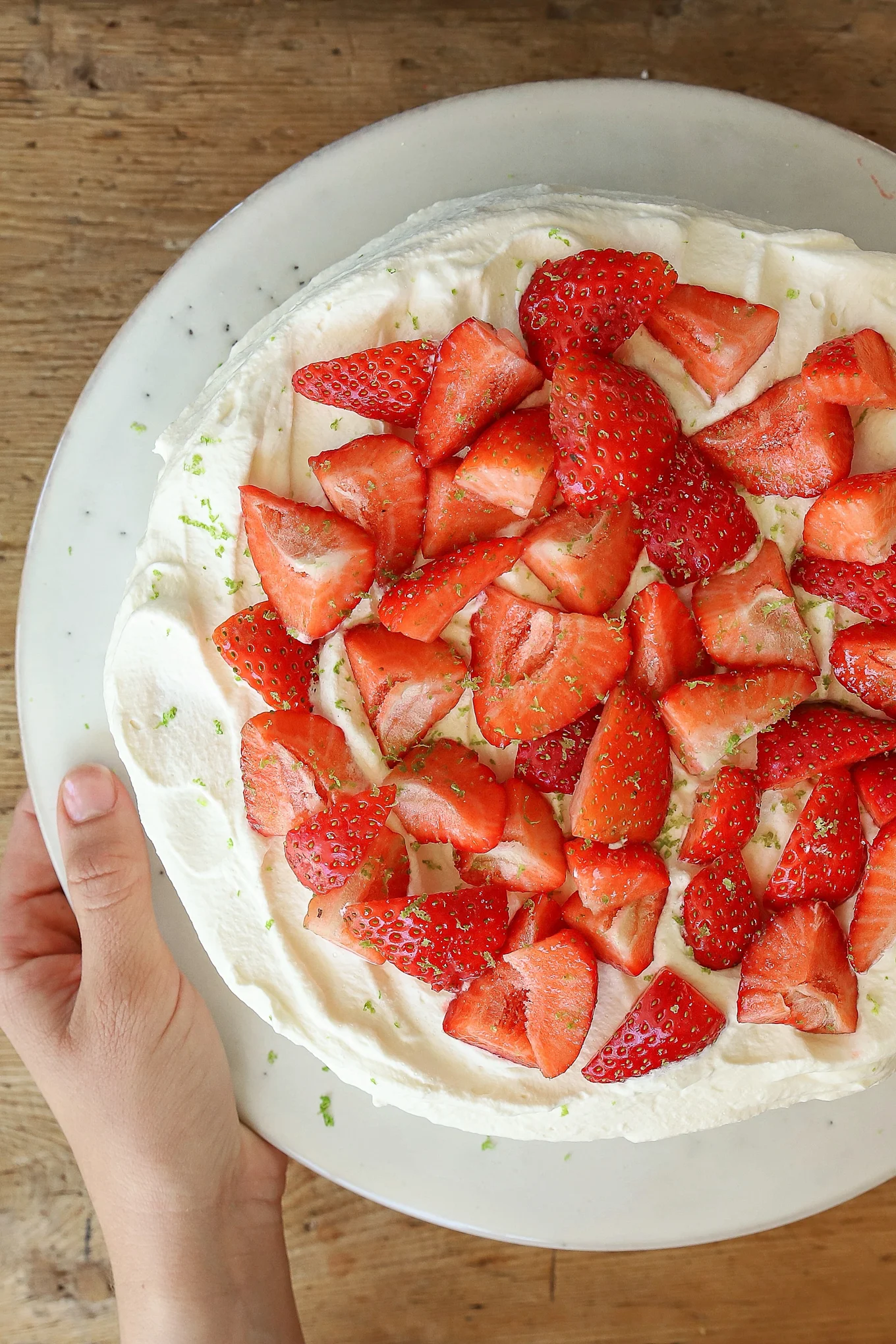Overhead shot of a hand holding a round cake with whipped cream, strawberries, and lime zest on a white stand.
