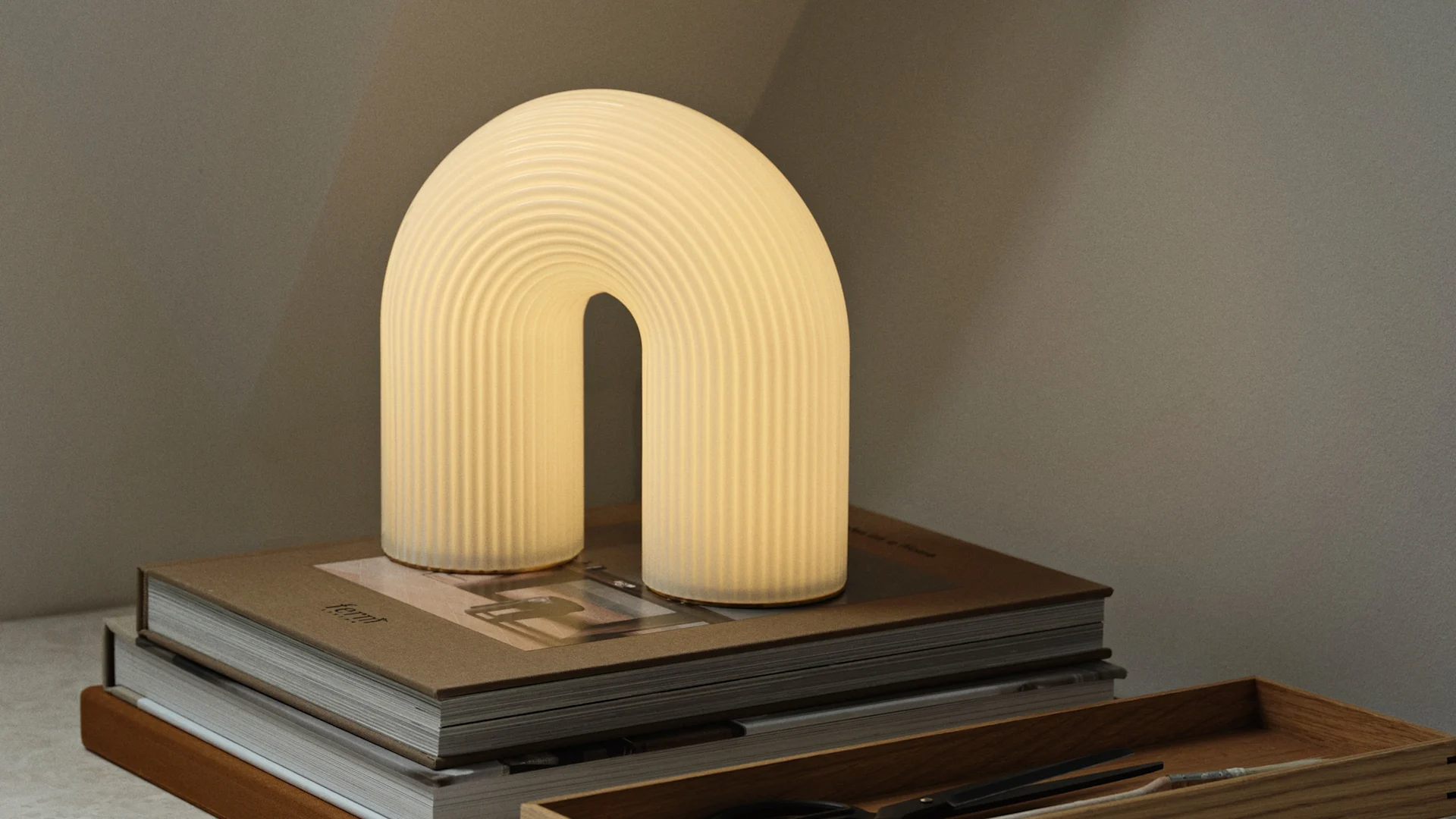 Warmly glowing, arch-shaped table lamp with a ribbed texture on a stack of brown books, with a wooden tray nearby.