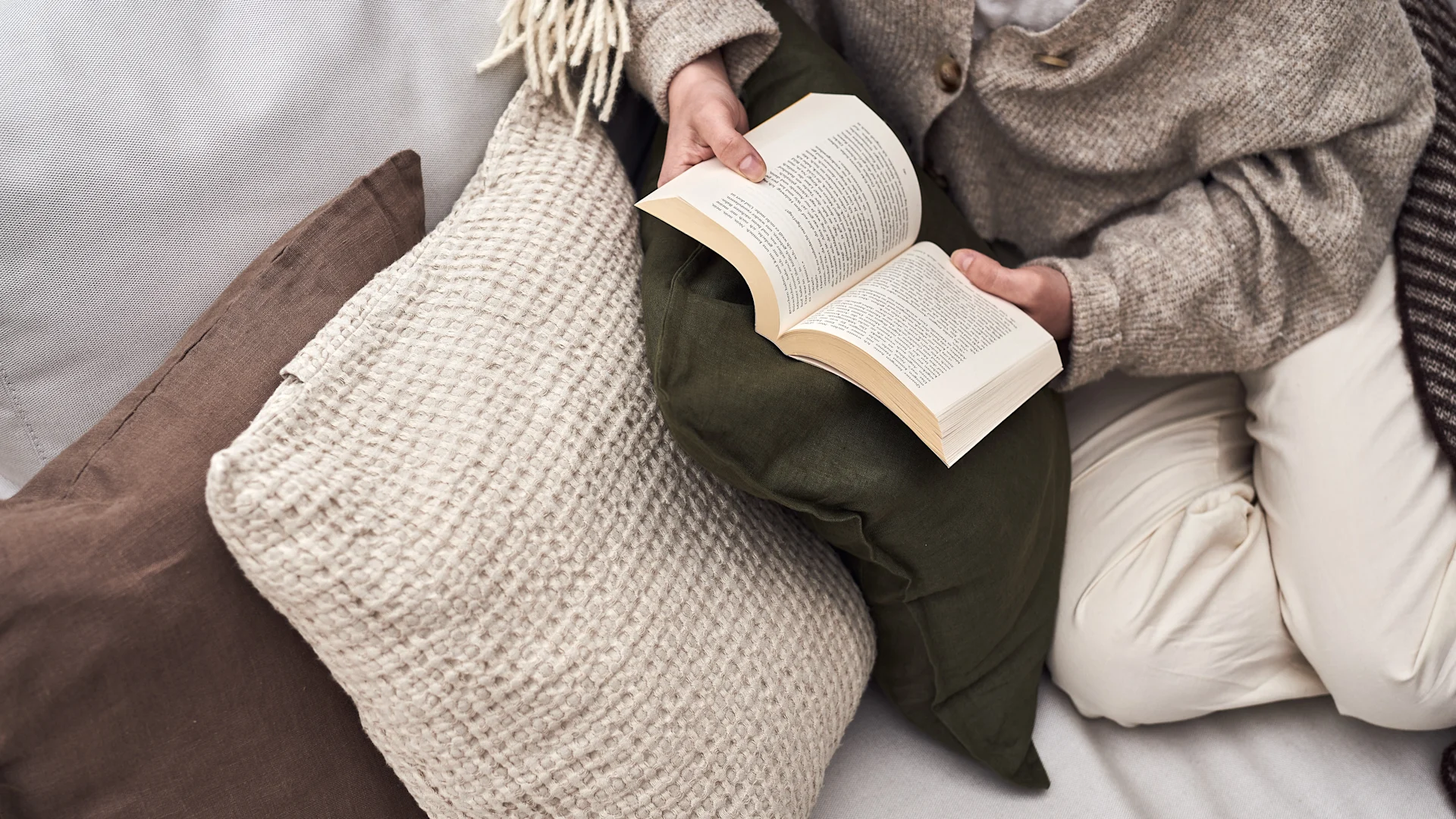 Person in a knit cardigan and light pants reading a book on a sofa with cozy pillows and a tasseled blanket.