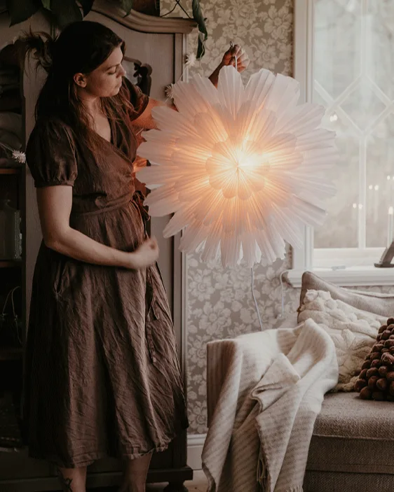 Woman in brown dress hangs an illuminated white paper star lantern in a cozy room.