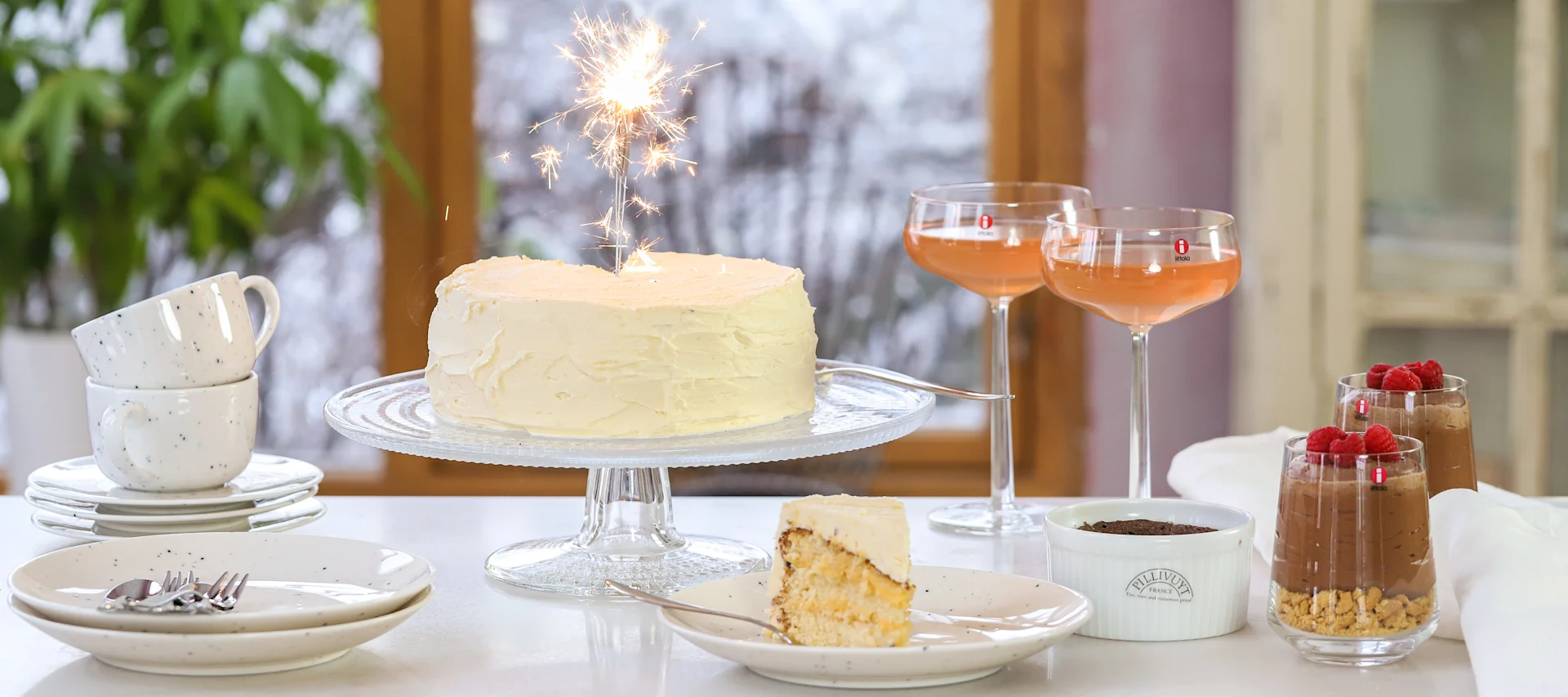 White frosted celebration cake with sparkler, slice of cake, pink drinks, chocolate mousse desserts on a table.