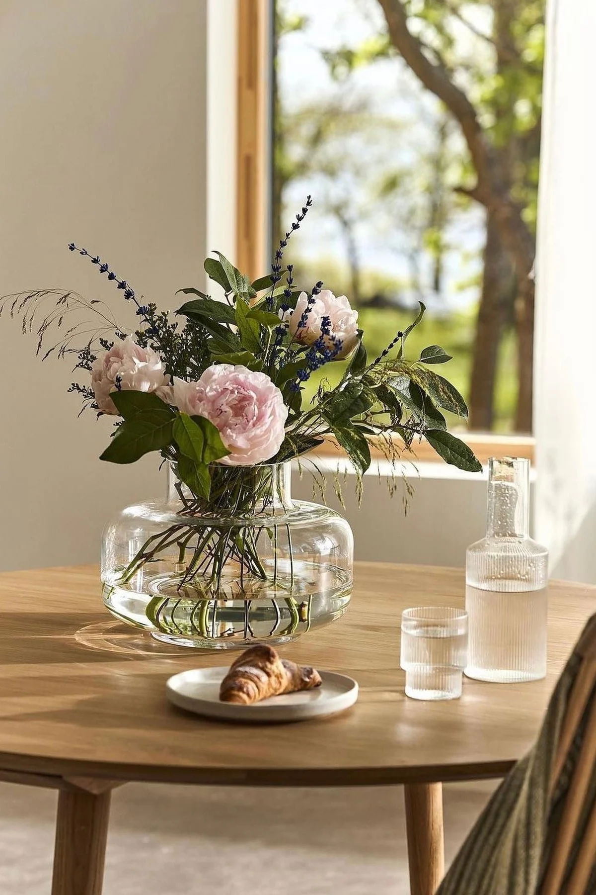 Clear glass vase with light pink peonies and lavender, a croissant, water carafe, and glass on a wooden table.
