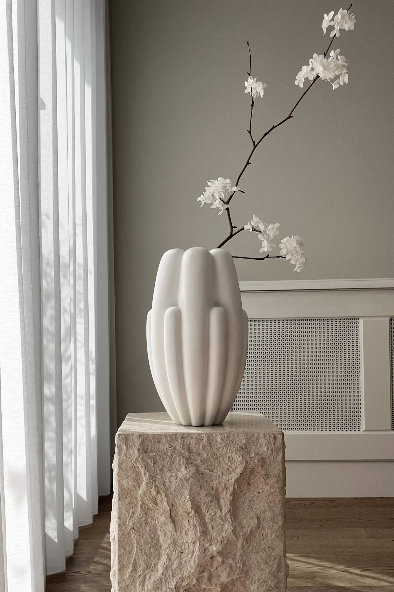 White fluted vase with white flowers on a textured stone pedestal, by a window with white blinds.