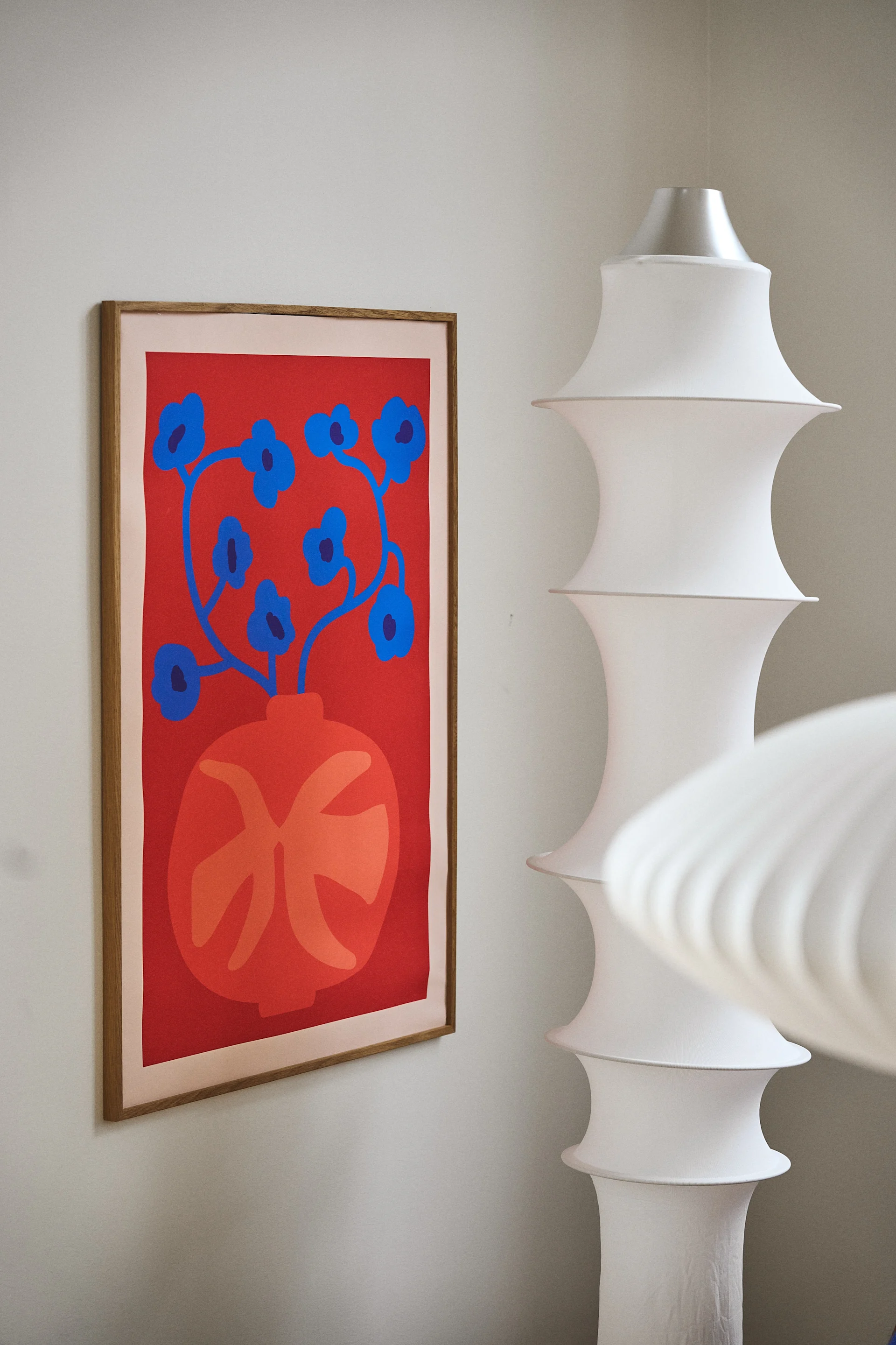 A framed abstract floral print with blue flowers in an orange vase on a red background, next to a tall, white, tiered lamp.