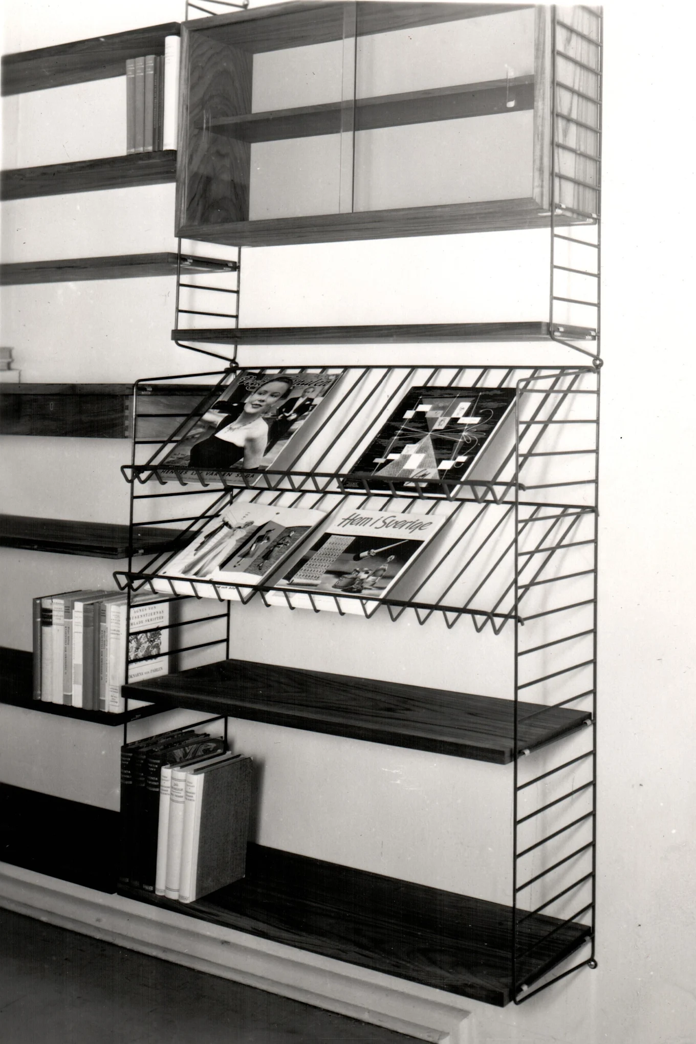 A wall-mounted shelving unit with dark wooden shelves, wire magazine racks, and a top cabinet with glass doors, all against a light wall.