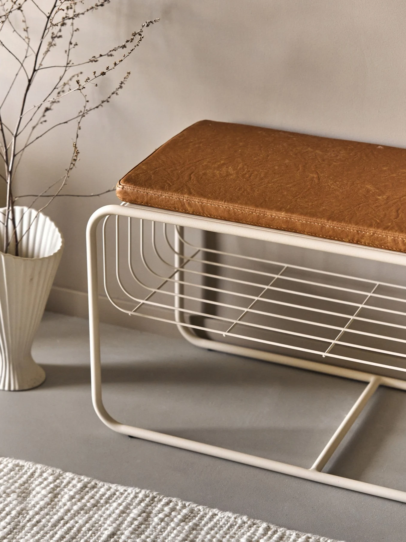 Modern beige metal bench with brown faux leather cushion, featuring an open slatted shelf. A white vase with branches and a rug are nearby.