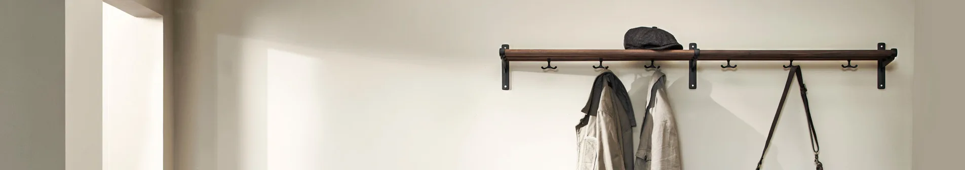 A wooden coat rack with black hooks on a light wall, holding a black cap, two jackets, and a dark bag.