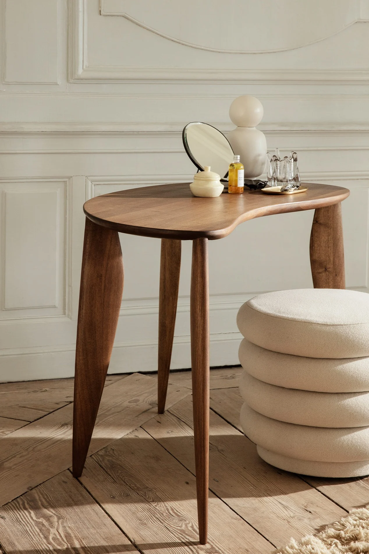 Organic-shaped wooden desk with mirror, oil, and decor, beside a stacked cream cushion stool against a paneled wall.