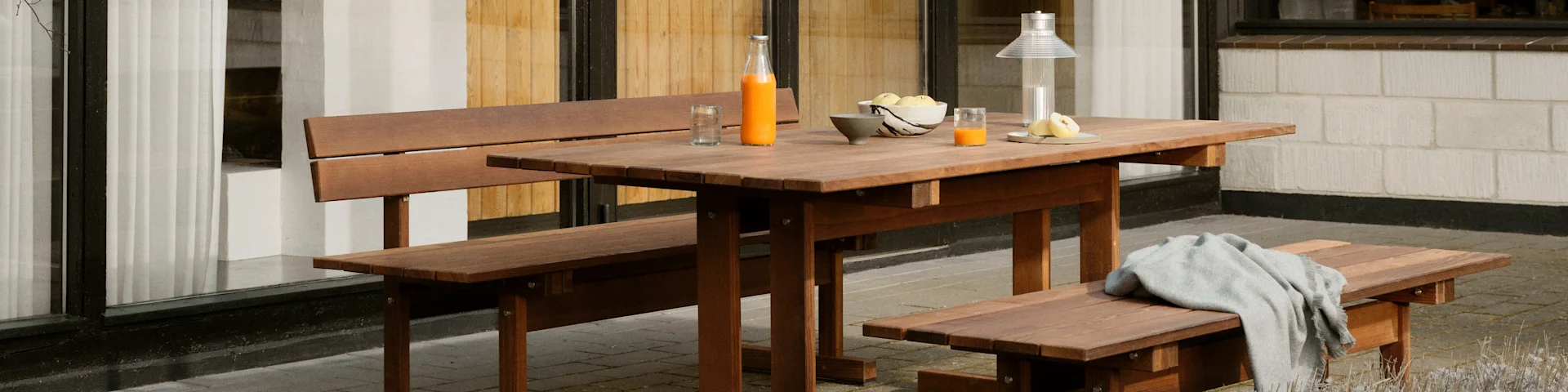 A wooden picnic table set with a bottle of juice and fruit on top, and a grey blanket on a bench.