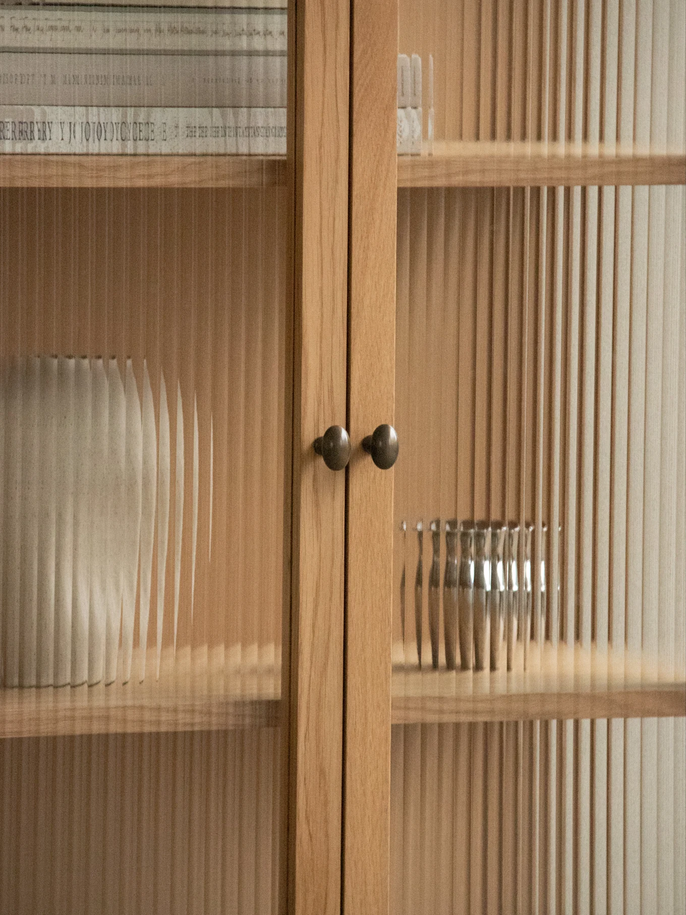Close-up of a light wood cabinet with reeded glass doors and round knobs, showing a textured vase and metal bowl inside.