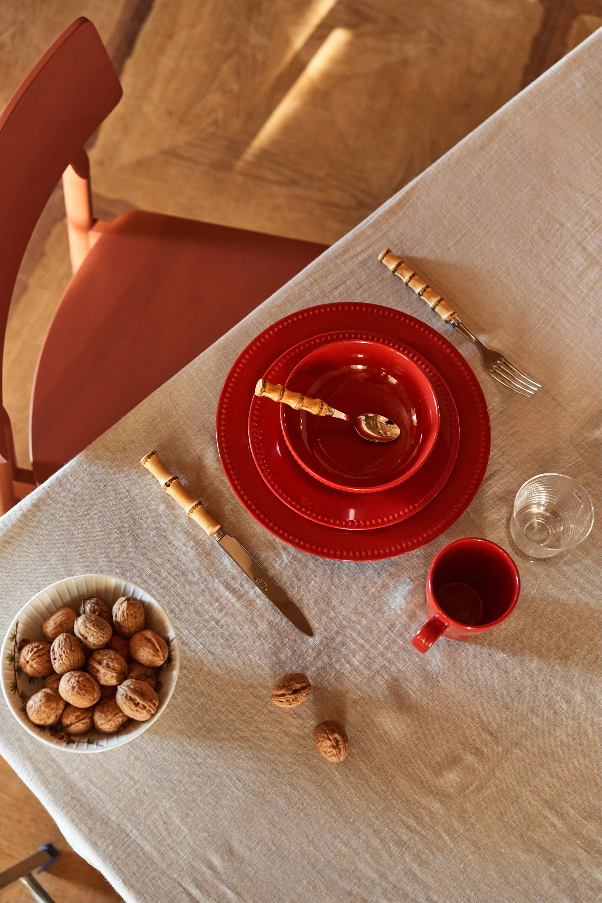 Dining table setting with red ceramic plates, bamboo cutlery, glass, mug, and a bowl of walnuts on a linen tablecloth.