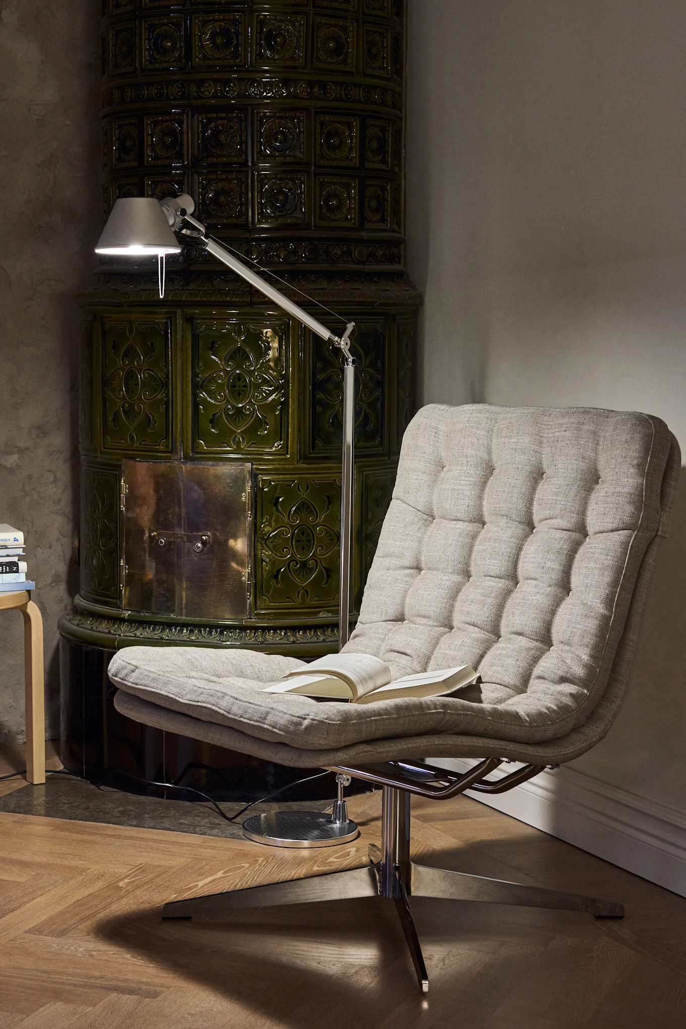 A cozy scene with a tufted lounge chair, an open book, and a floor lamp beside a decorative green tiled stove.