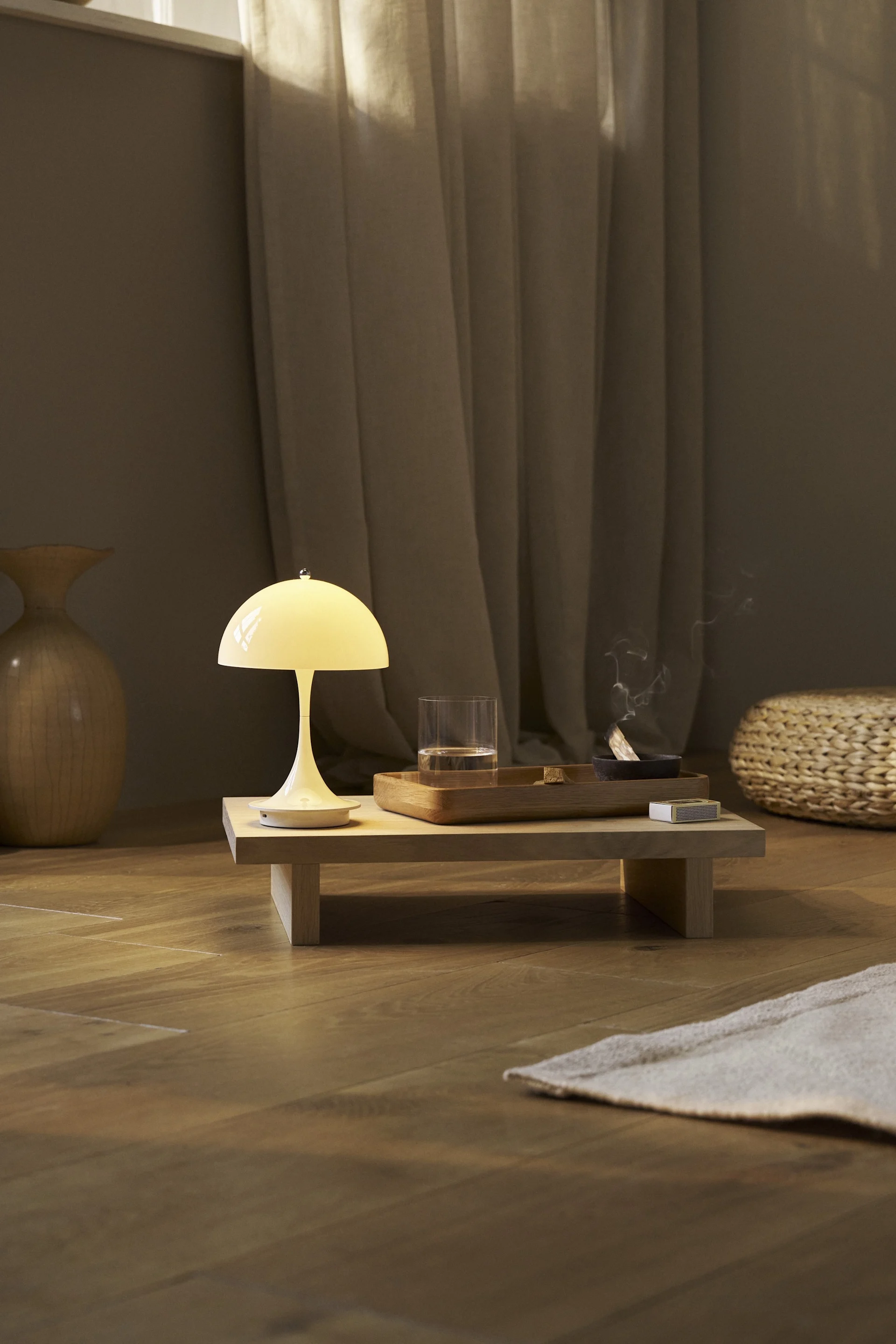 Cream mushroom lamp on a low wooden table with a glass of water, burning incense, and matches in a cozy, dimly lit room.