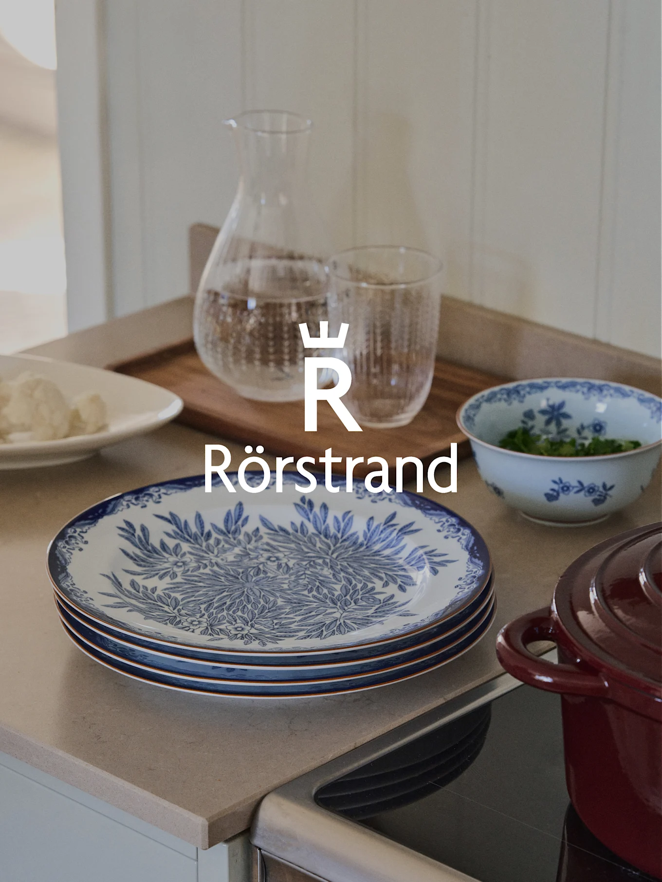 Rörstrand logo over kitchen counter with blue and white plates, carafe, glass, and red pot.