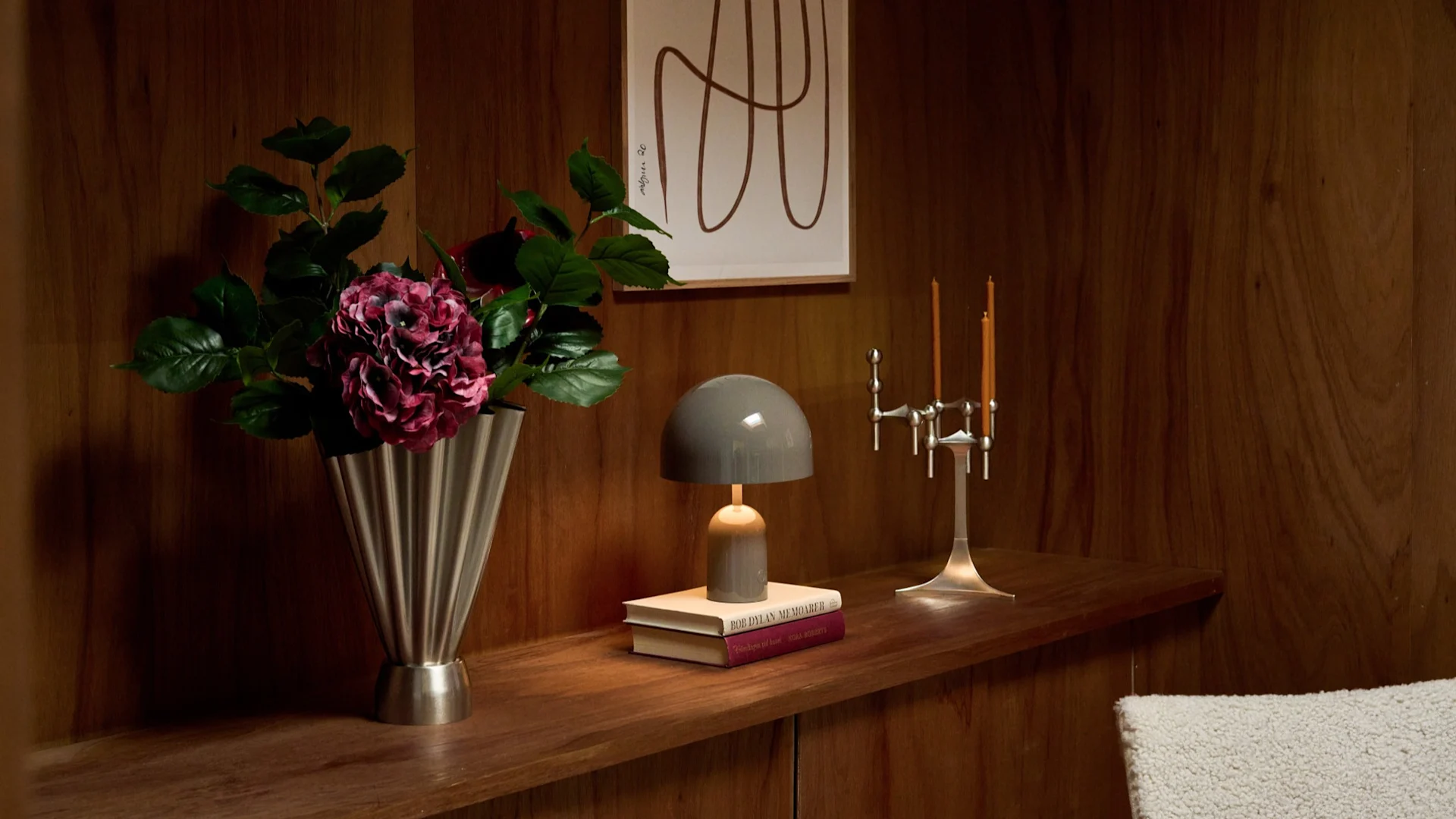Vase with dark pink flowers, grey mushroom lamp on books, silver candelabra on a wooden shelf with art.
