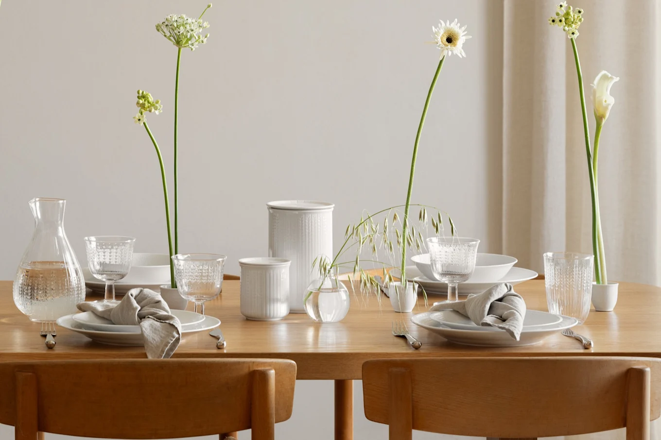 Elegant dining table set for two with white plates, linen napkins, patterned glass drinkware, and a variety of white flowers in vases.
