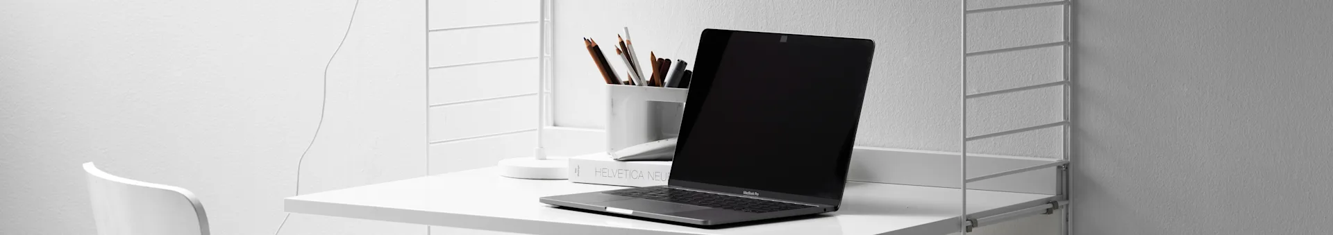 A silver laptop, a white cup of pencils, and a book sit on a minimalist white desk shelf against a white wall.