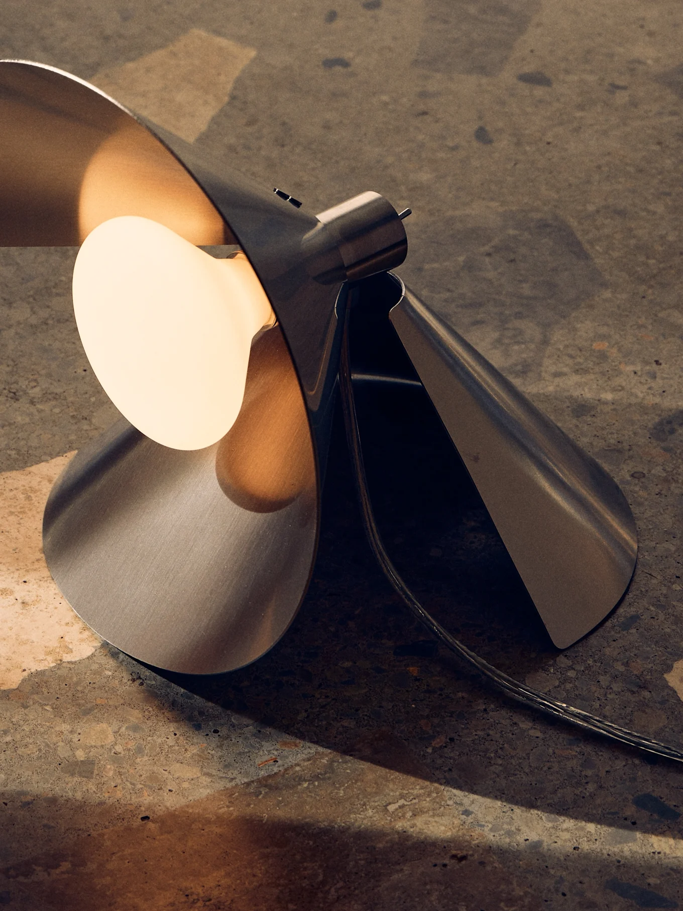 Modern brushed metal table lamp with a sculptural, folded design and a warm, exposed bulb on a textured surface.