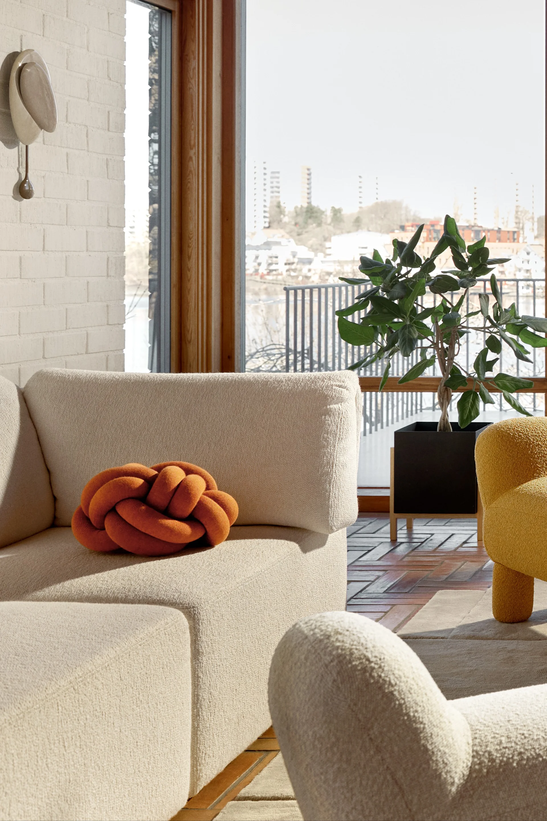 A cozy living room with an off-white sofa, an orange knot pillow, and a yellow armchair, overlooking a city and water view.