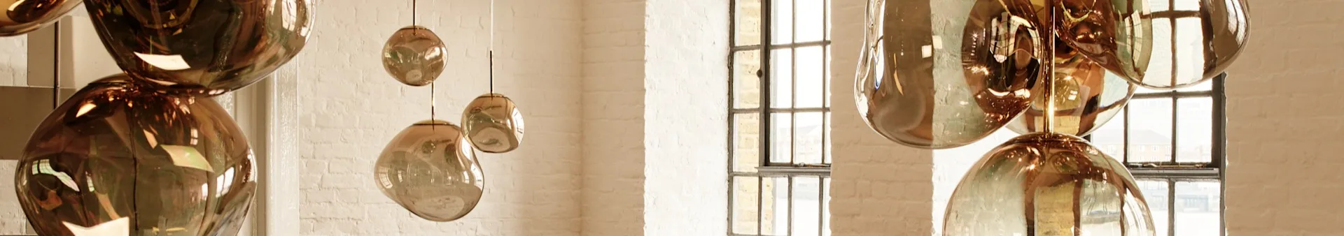Metallic copper-colored, irregularly shaped pendant lights hang against a white brick wall with windows in the background.