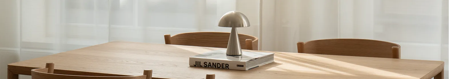 Light wood dining table and four chairs. Silver mushroom lamp and book on table. Sunlight through sheer white curtains.