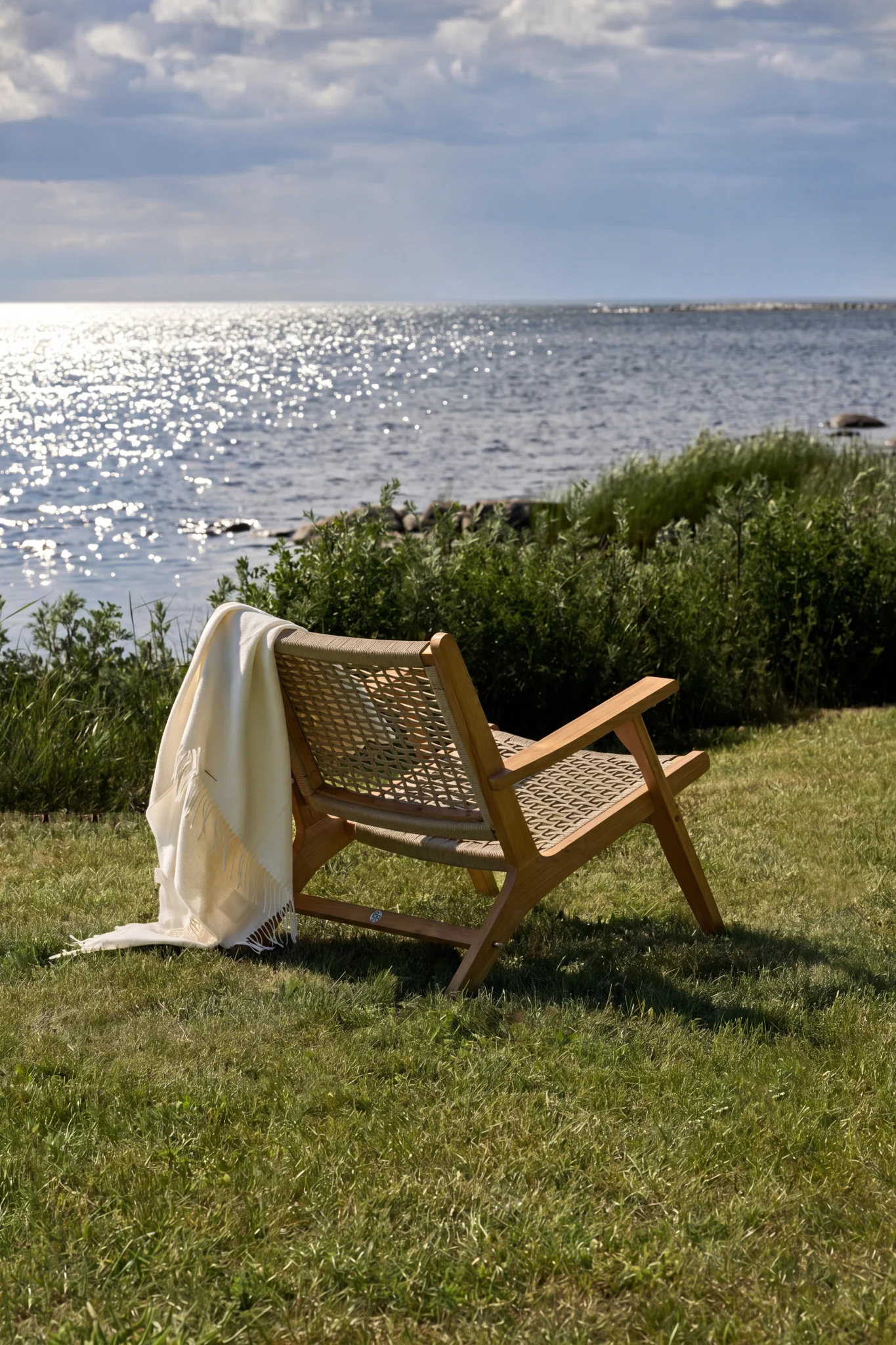 Wooden lounge chair with a woven seat and back, draped with a white blanket, facing a sparkling ocean under a cloudy sky.