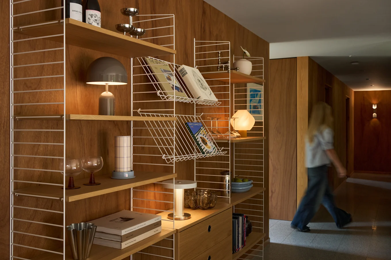 A blonde person walks down a wooden hallway next to a modern shelving unit filled with books, lamps, and decorative items.