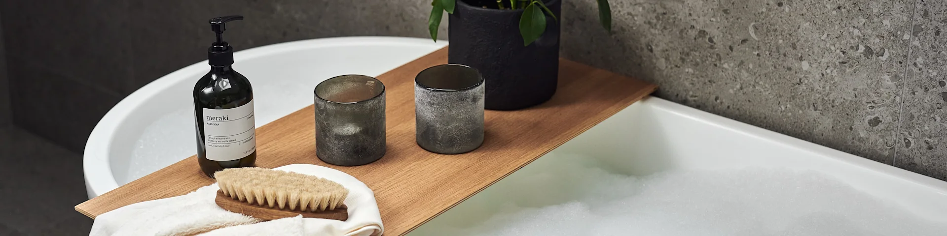 A bath caddy with a pump bottle, two grey candle holders, and a body brush on a towel across a bubble-filled bathtub.