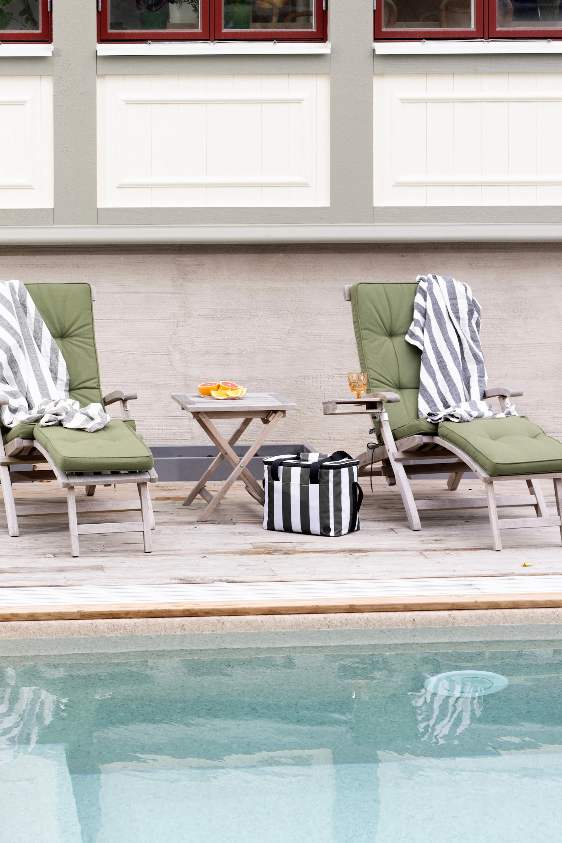 Two wooden lounge chairs with green cushions and striped blankets on a wooden deck next to a pool. A cooler bag and grapefruit are nearby.